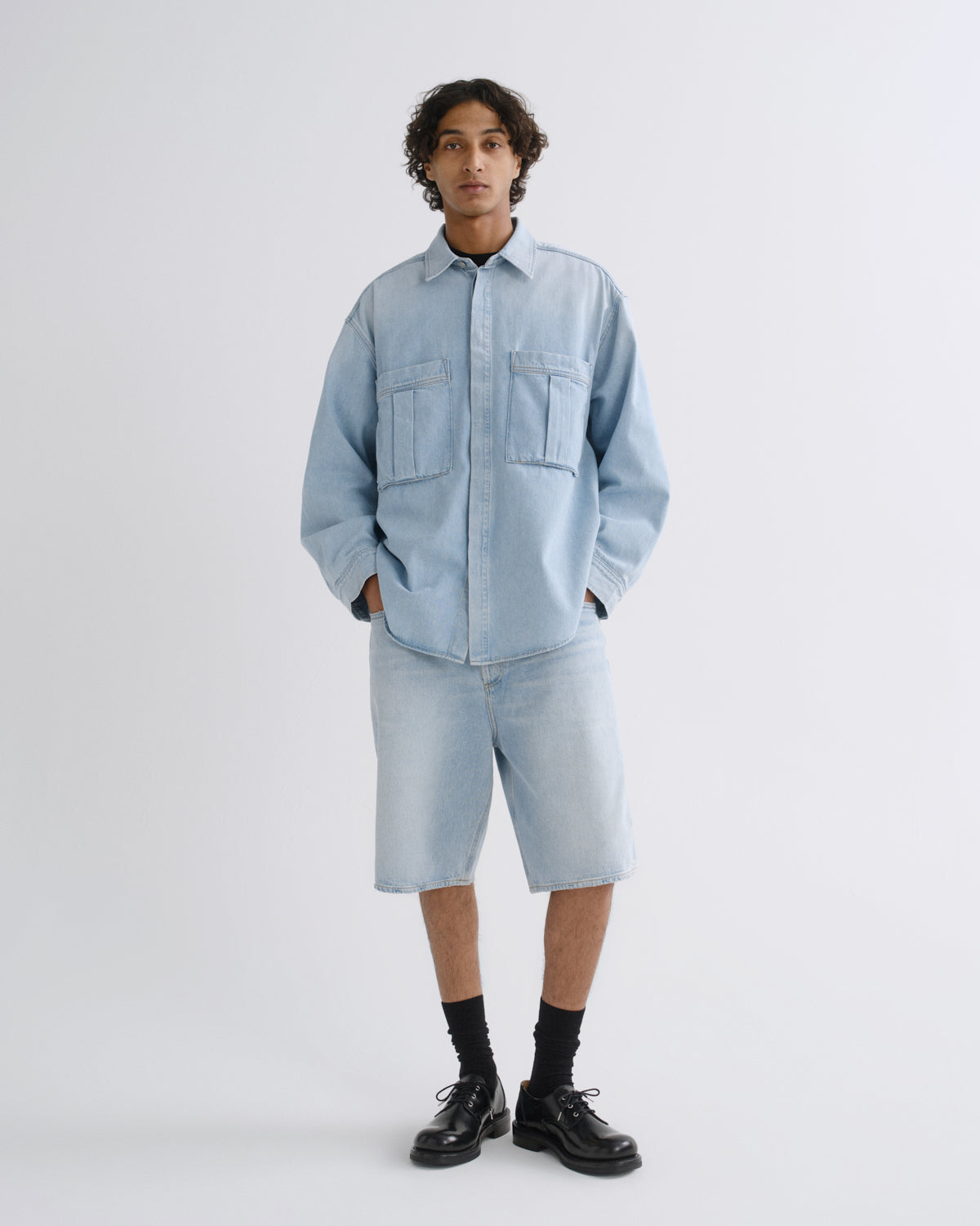 A model stands against a light gray studio background wearing a light blue denim shirt-jacket and matching denim shorts, paired with black socks and black leather shoes.