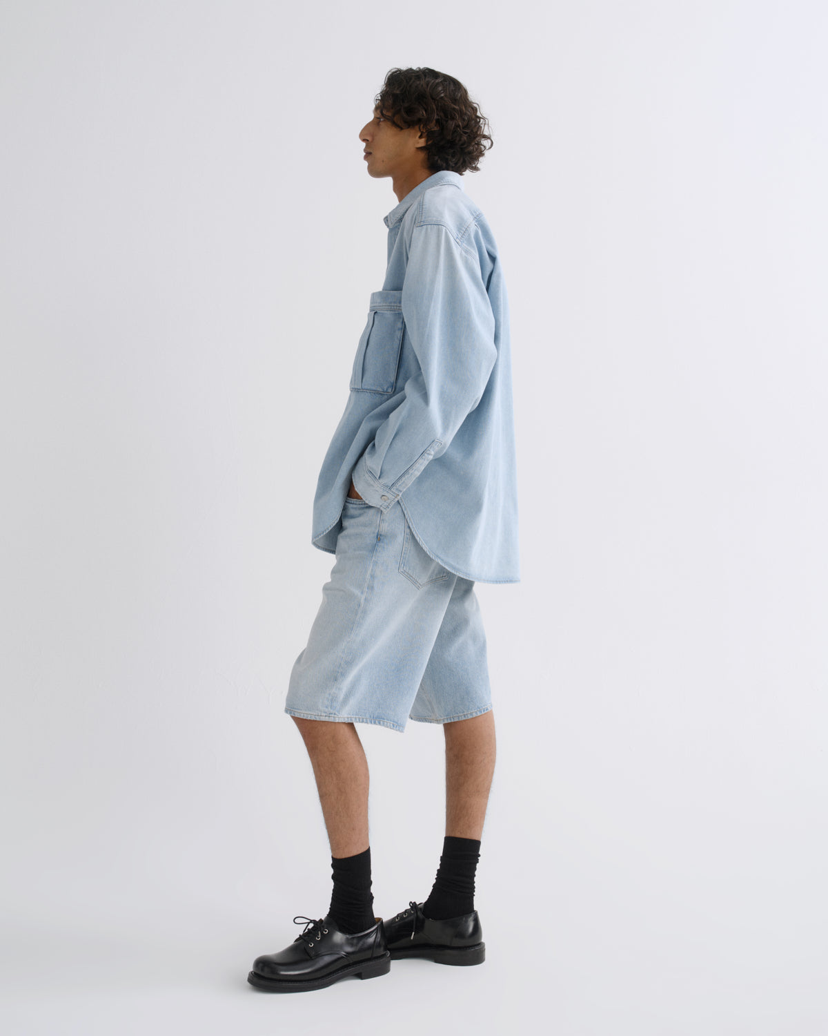A model is shown in profile wearing a light blue denim shirt and matching denim shorts, paired with black socks and black leather shoes, against a plain white studio background.