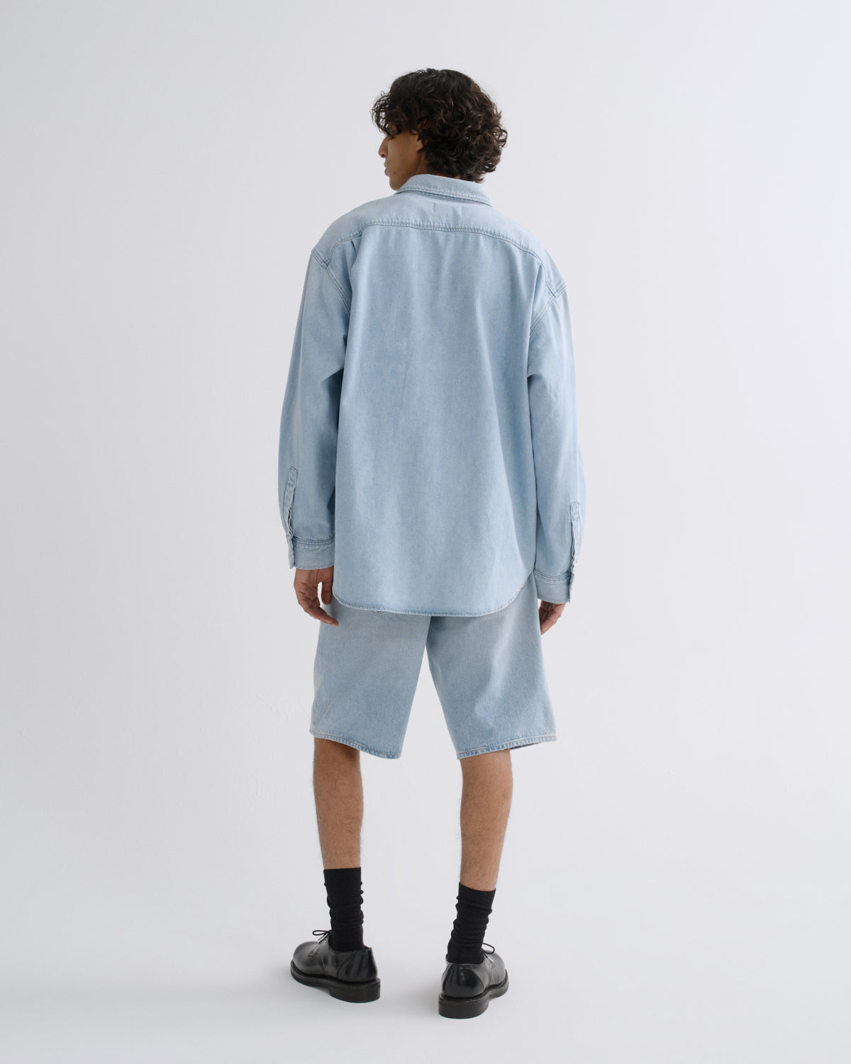A model shown from the back wearing a light blue denim shirt and matching denim shorts, paired with black socks and black leather shoes, against a plain light gray studio background.