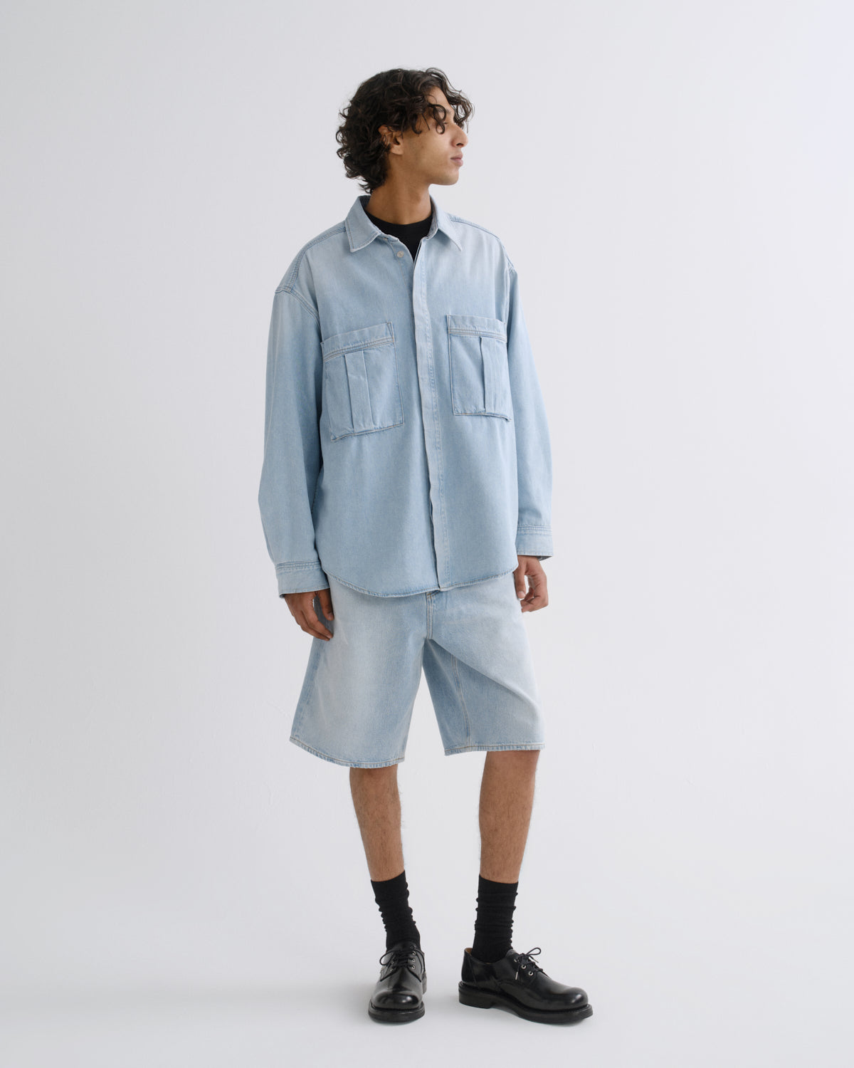 A model wearing a light blue oversized denim shirt and matching light blue denim shorts, styled with black crew socks and black leather shoes, standing against a plain light gray studio background.