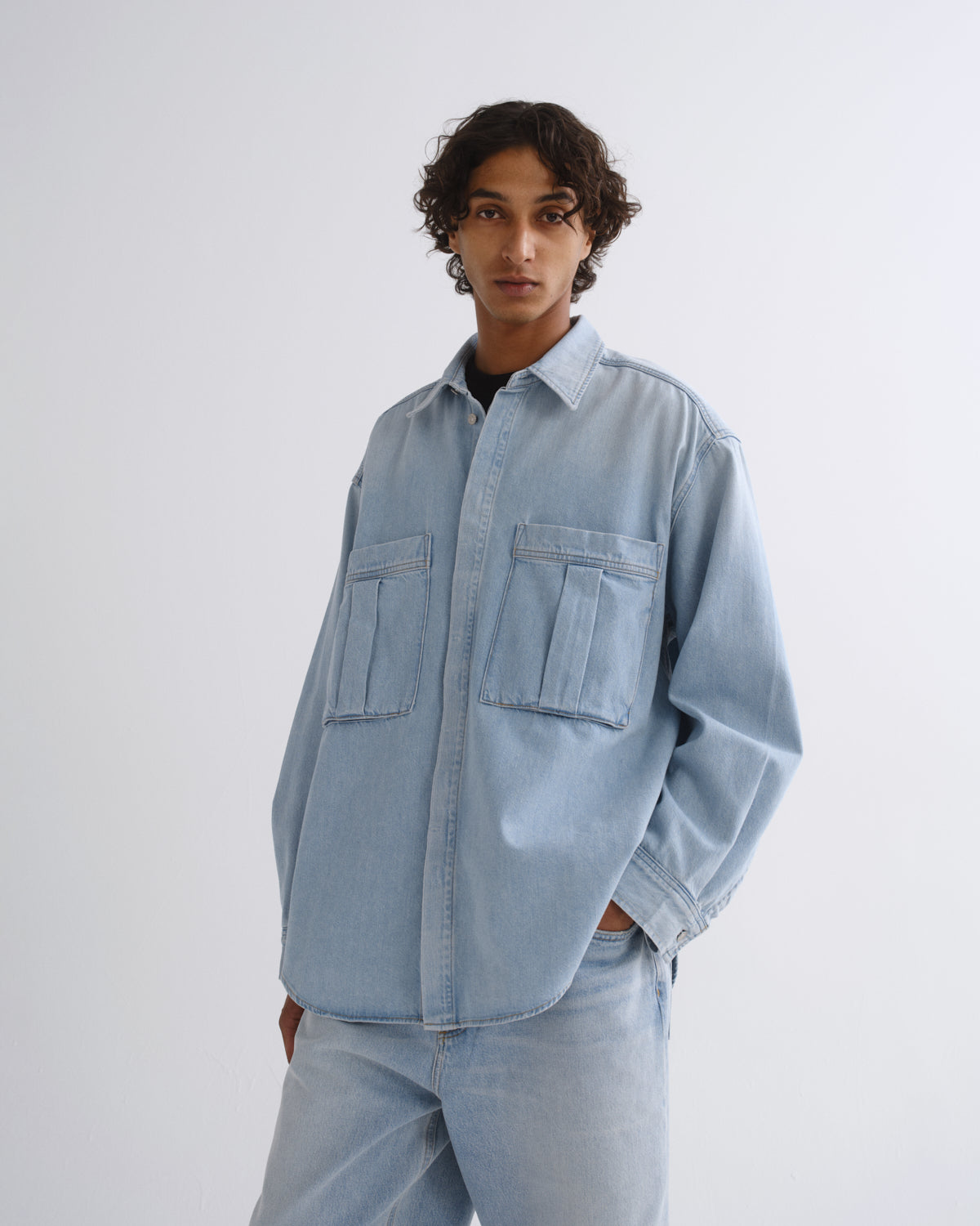 A model wearing a light blue denim button-up shirt and matching light blue jeans, standing against a plain light gray studio background.