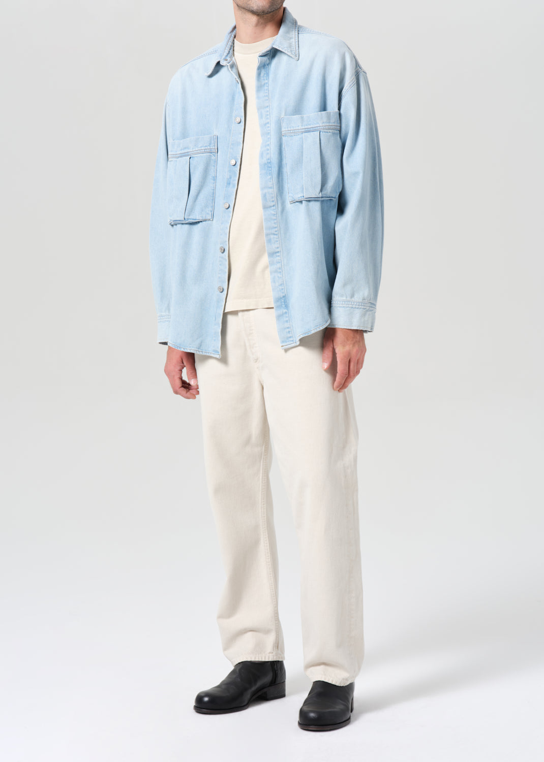 Person wearing a light blue denim shirt over a white t-shirt, paired with white pants and black shoes on a white background.