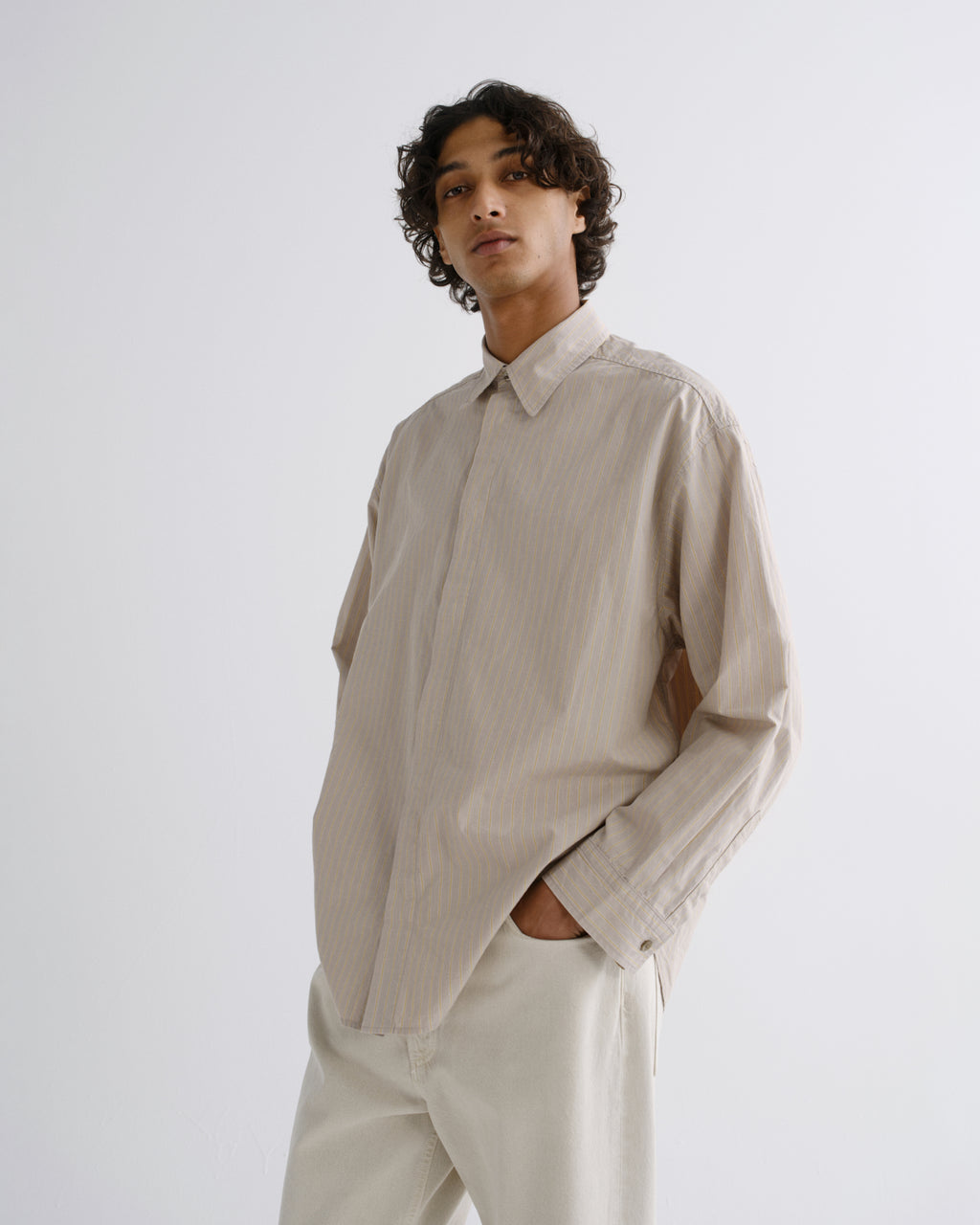 A man wearing a light beige long-sleeve linen shirt and matching beige trousers, standing against a plain light gray studio background.