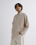 A man wearing a light beige long-sleeve linen shirt and matching beige trousers, standing against a plain light gray studio background.