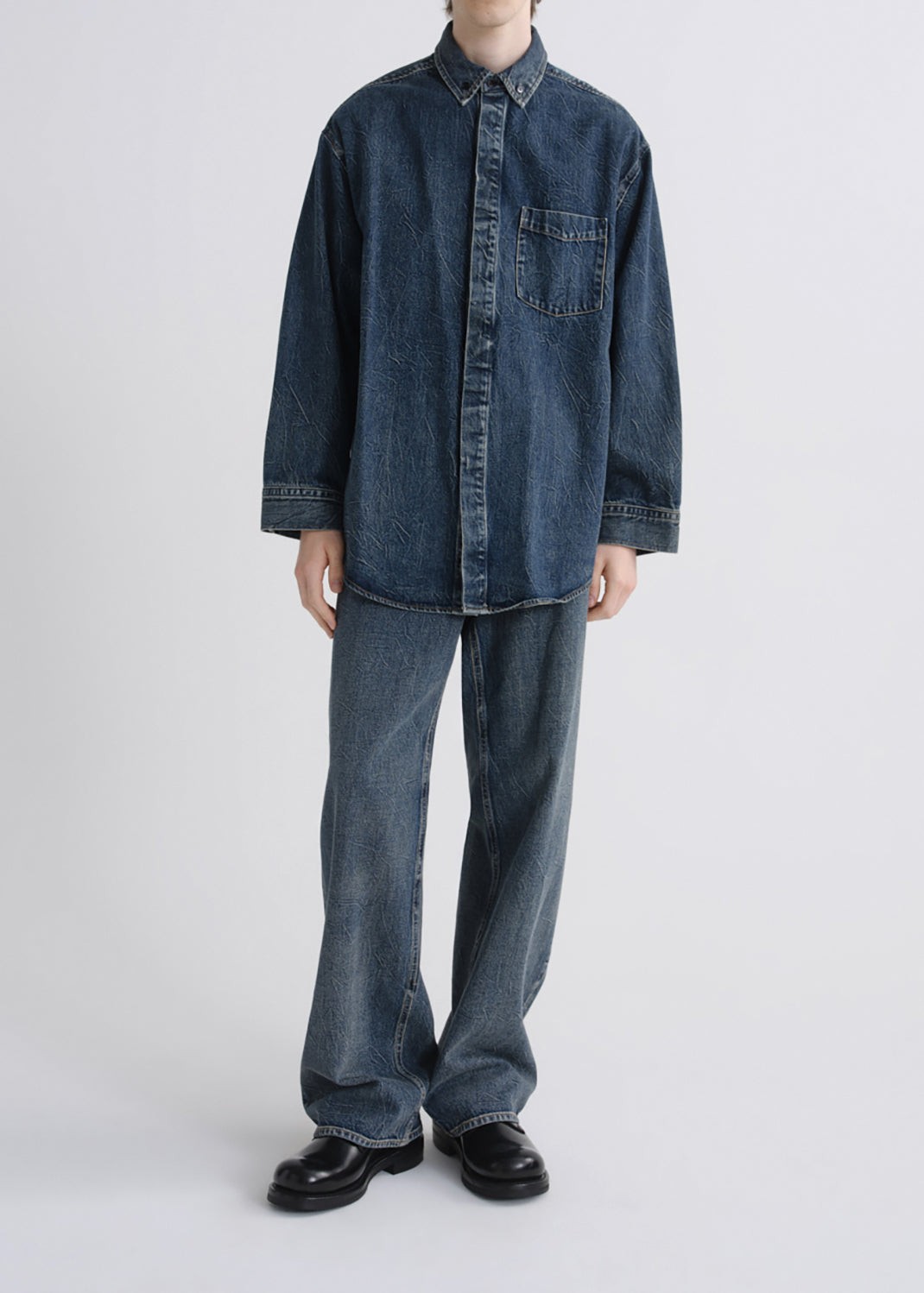 A model wearing an oversized dark blue denim button-up shirt and matching wide-leg jeans, paired with black leather shoes, against a grey background.