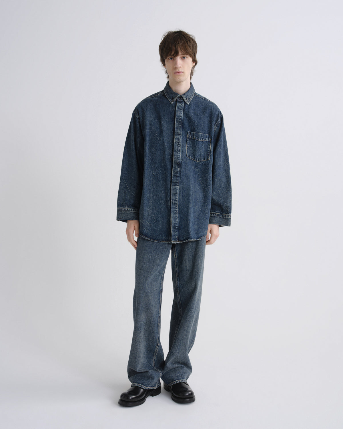 A model stands against a light gray studio background wearing an oversized dark blue denim button-up shirt with matching wide-leg denim jeans and black leather shoes.