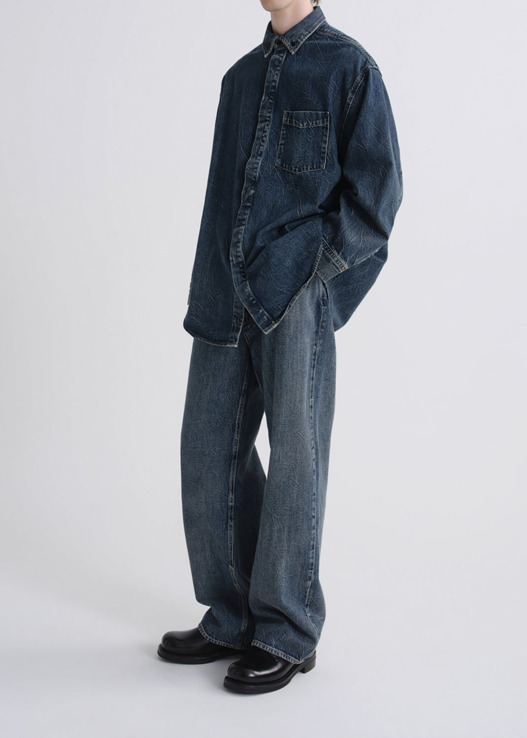 A model wearing an oversized dark blue denim shirt and matching wide-leg denim pants, paired with black leather shoes, photographed against a grey background.