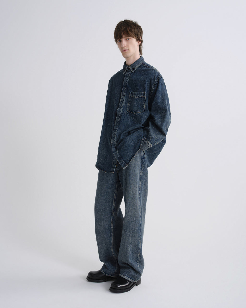 A model wearing an oversized dark blue denim shirt and matching wide-leg denim pants, standing against a plain light gray studio background.