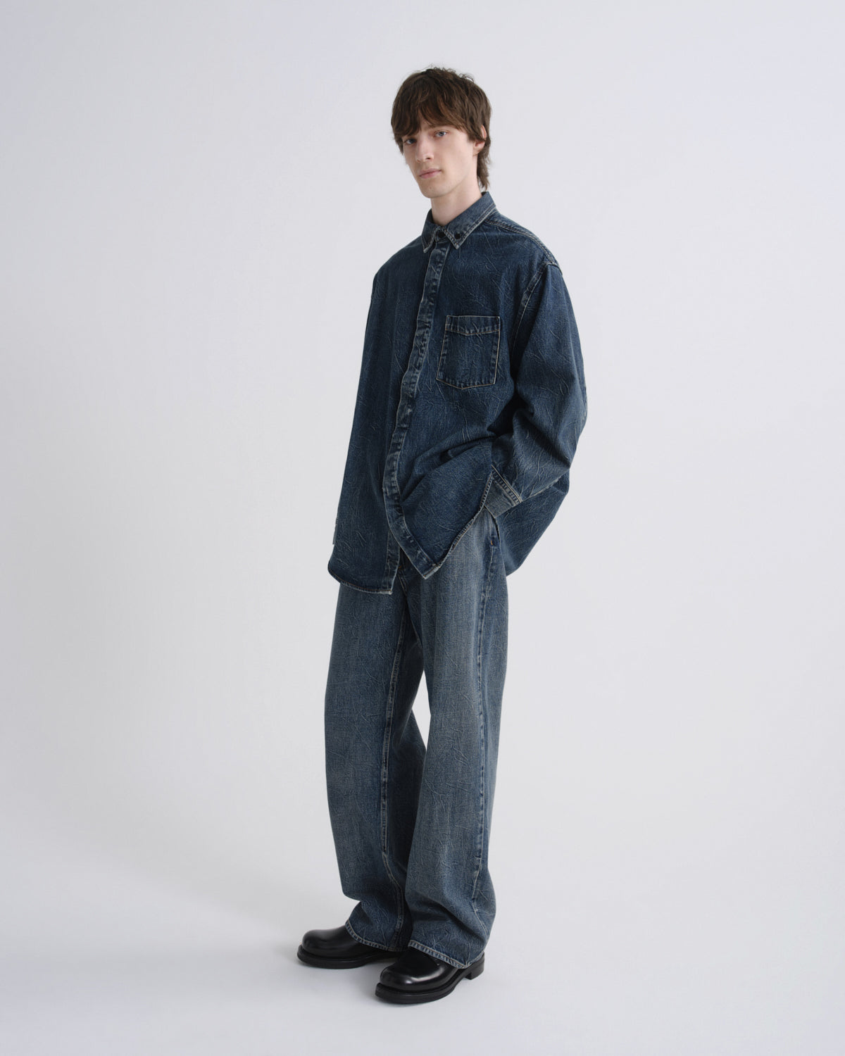 A model wearing an oversized dark blue denim shirt and matching wide-leg denim pants, standing against a plain light gray studio background.
