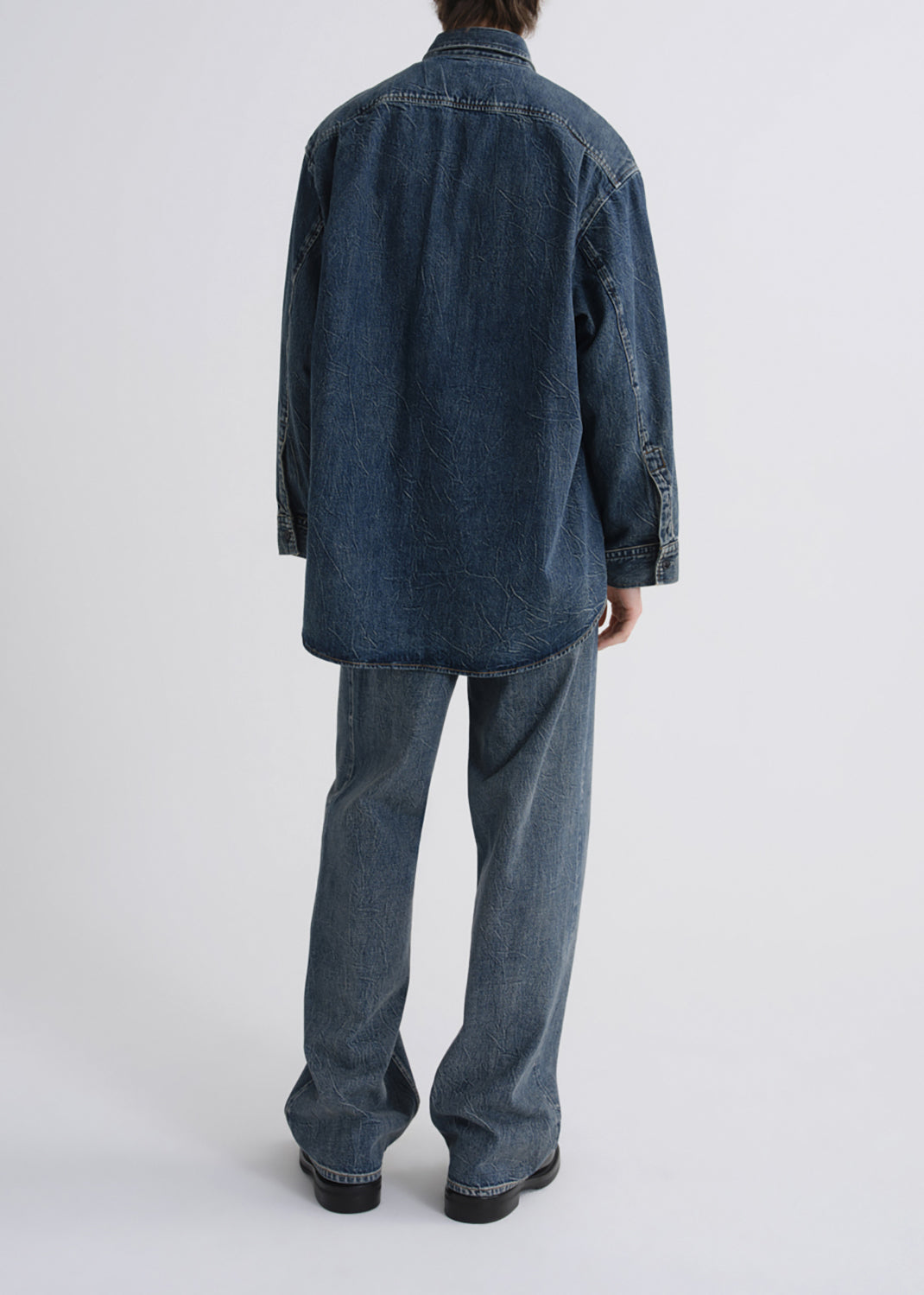 A model viewed from the back wearing an oversized dark blue denim shirt and matching wide-leg denim jeans against a grey background.