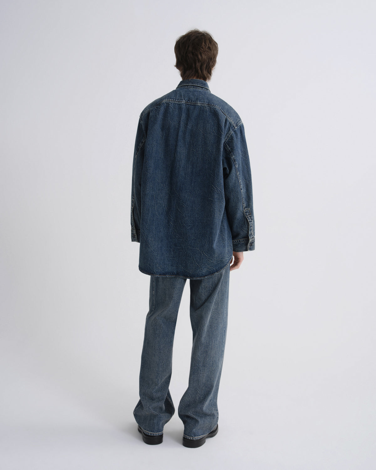A model viewed from the back wearing an oversized dark blue denim shirt and matching wide-leg jeans against a light gray studio background.
