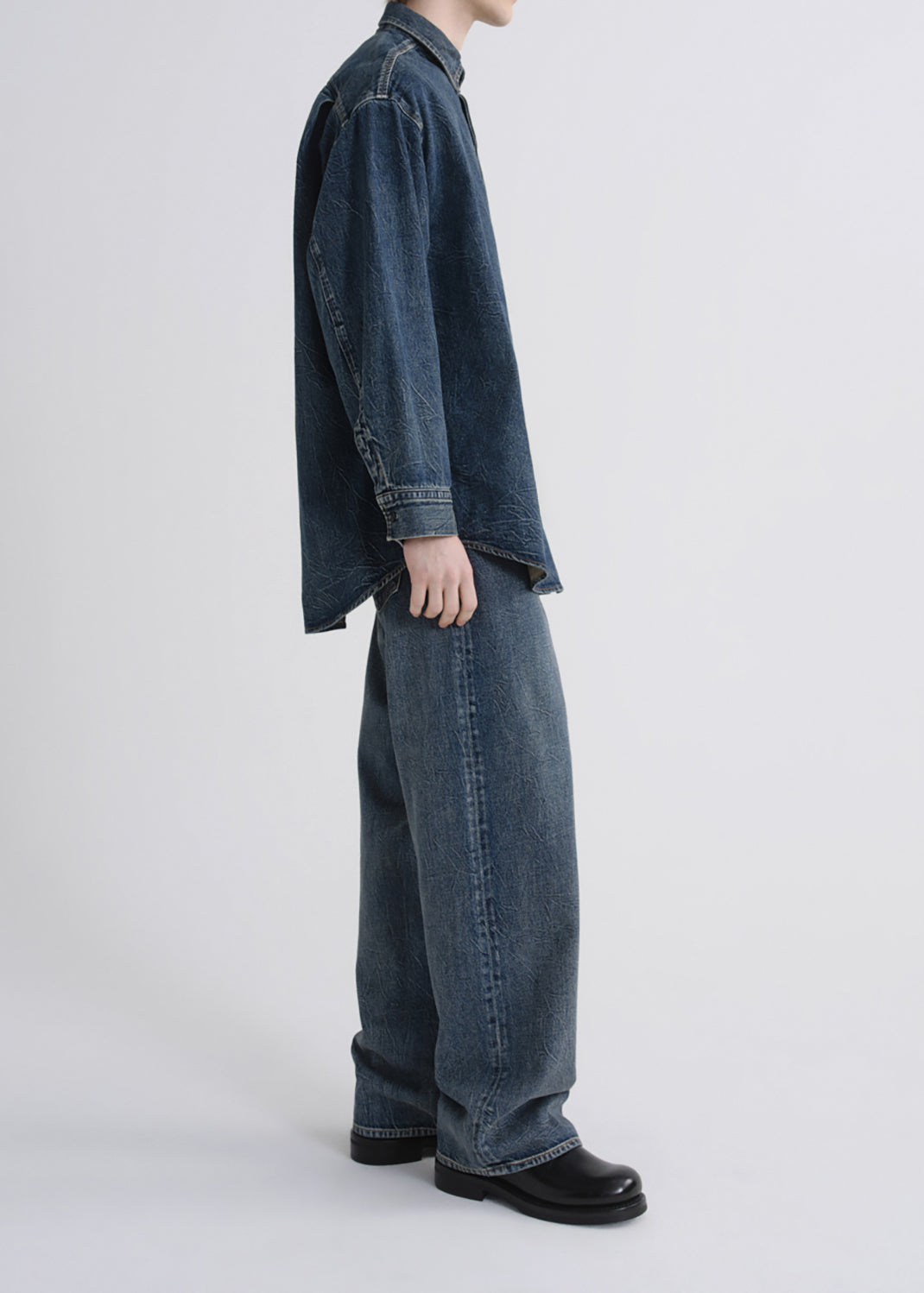 A side-profile studio shot of a model wearing a dark blue indigo denim shirt and matching wide-leg jeans, paired with black leather shoes, against a grey background.