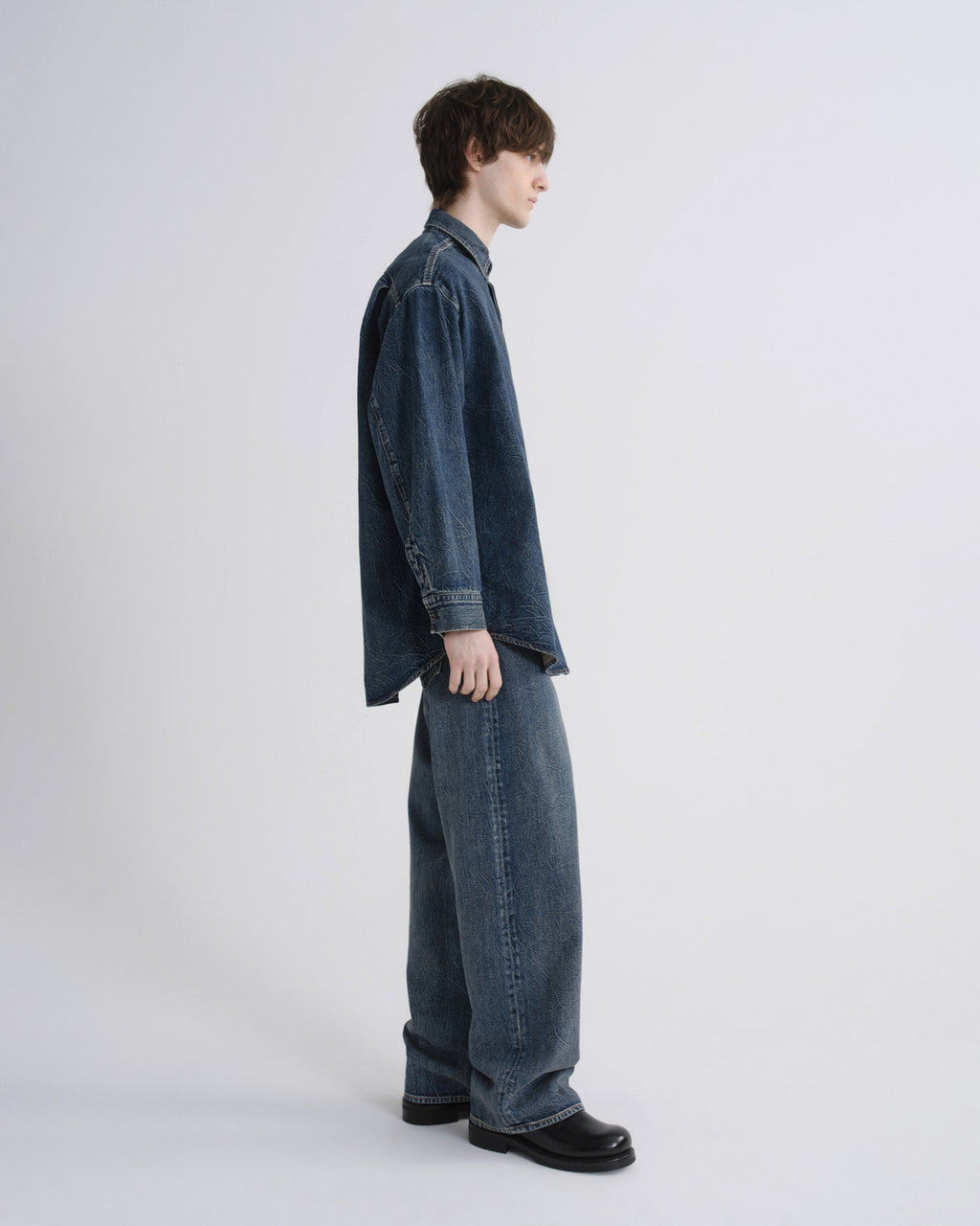 A model shown in profile wearing a dark blue denim shirt and matching wide-leg denim jeans, paired with black leather shoes, against a plain light gray studio background.