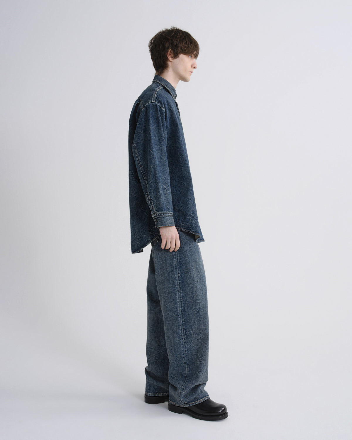 A model shown in profile wearing a dark blue denim shirt and matching wide-leg denim jeans, paired with black leather shoes, against a plain light gray studio background.