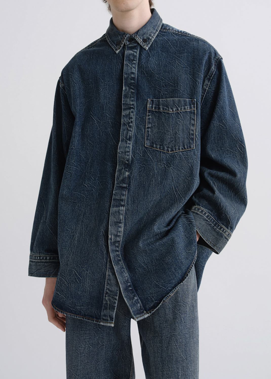 A person wearing an oversized dark indigo denim button-up shirt with a chest pocket and matching dark denim pants, photographed against a grey background.