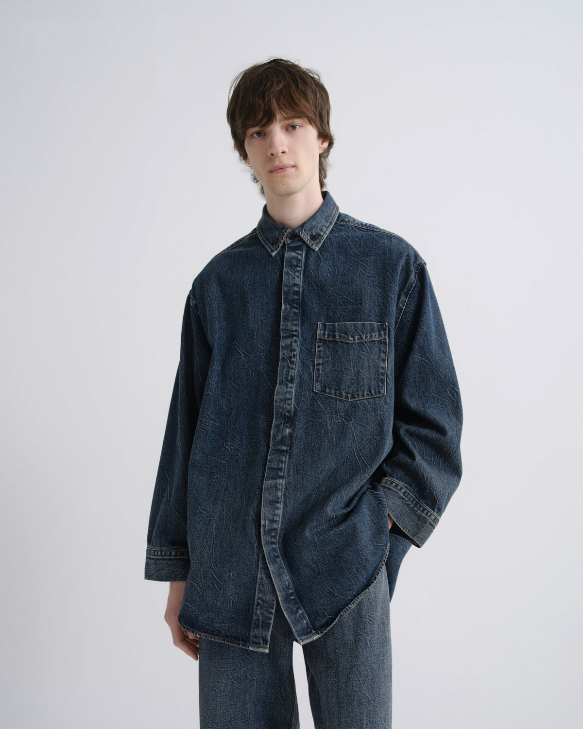 A man wearing an oversized dark indigo denim button-up shirt and matching jeans, standing against a plain light gray studio background.