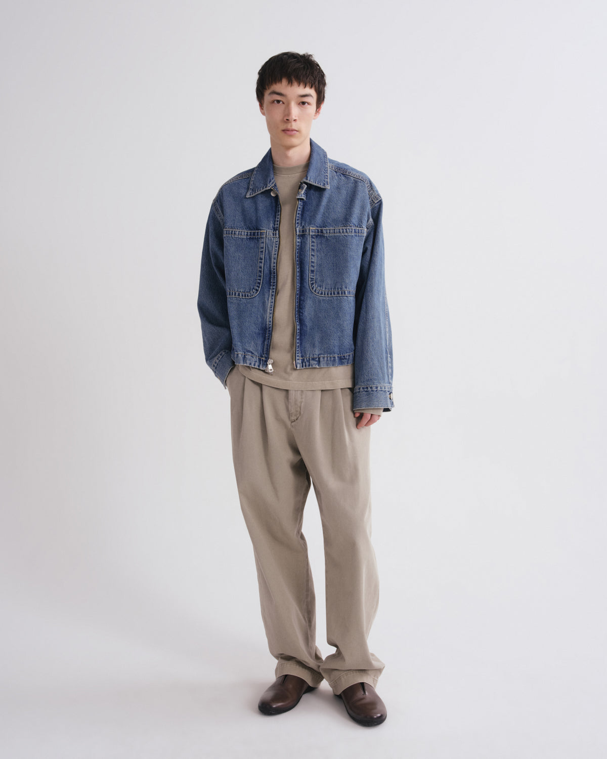 A model stands against a light gray studio background wearing a blue denim jacket over a beige knit top, paired with beige trousers and dark brown loafers.