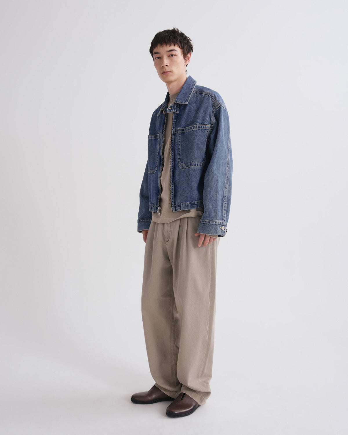 A male model stands against a light gray studio background wearing a blue denim jacket over a beige shirt and beige wide-leg trousers, with dark brown shoes.