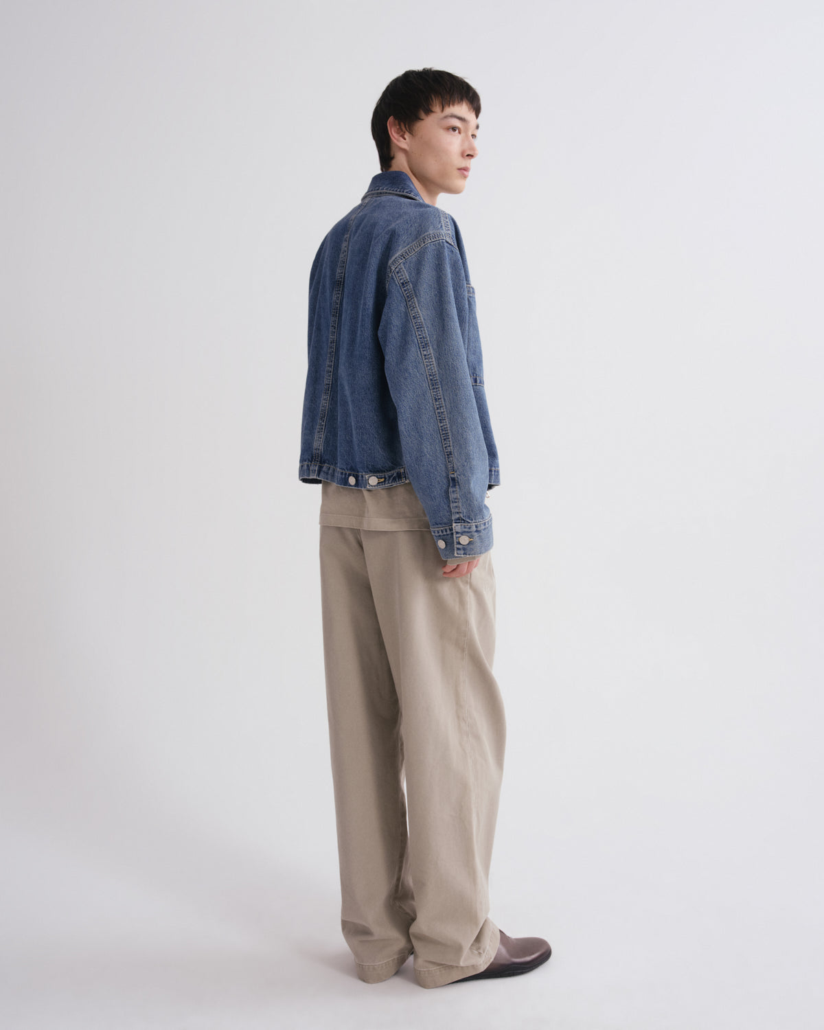 A model standing in profile wearing a cropped blue denim jacket, beige wide-leg trousers, and dark brown loafers against a plain light gray studio background.