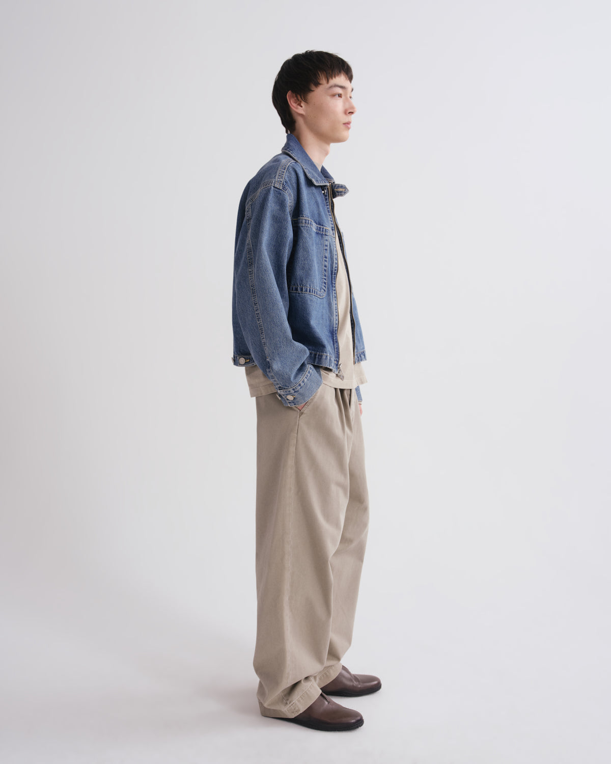 A model standing in profile wearing a blue denim jacket over a beige shirt, paired with beige trousers and dark brown shoes, against a plain light gray studio background.