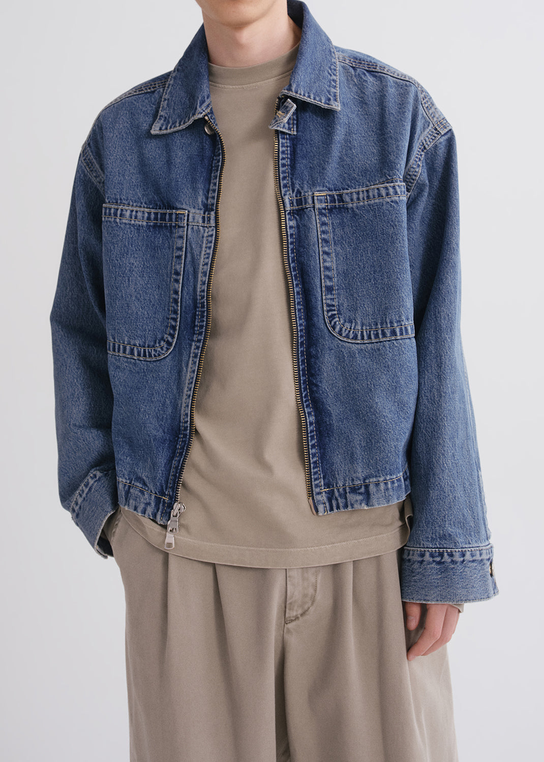 Person wearing a blue denim jacket over a beige shirt and pants on a grey background