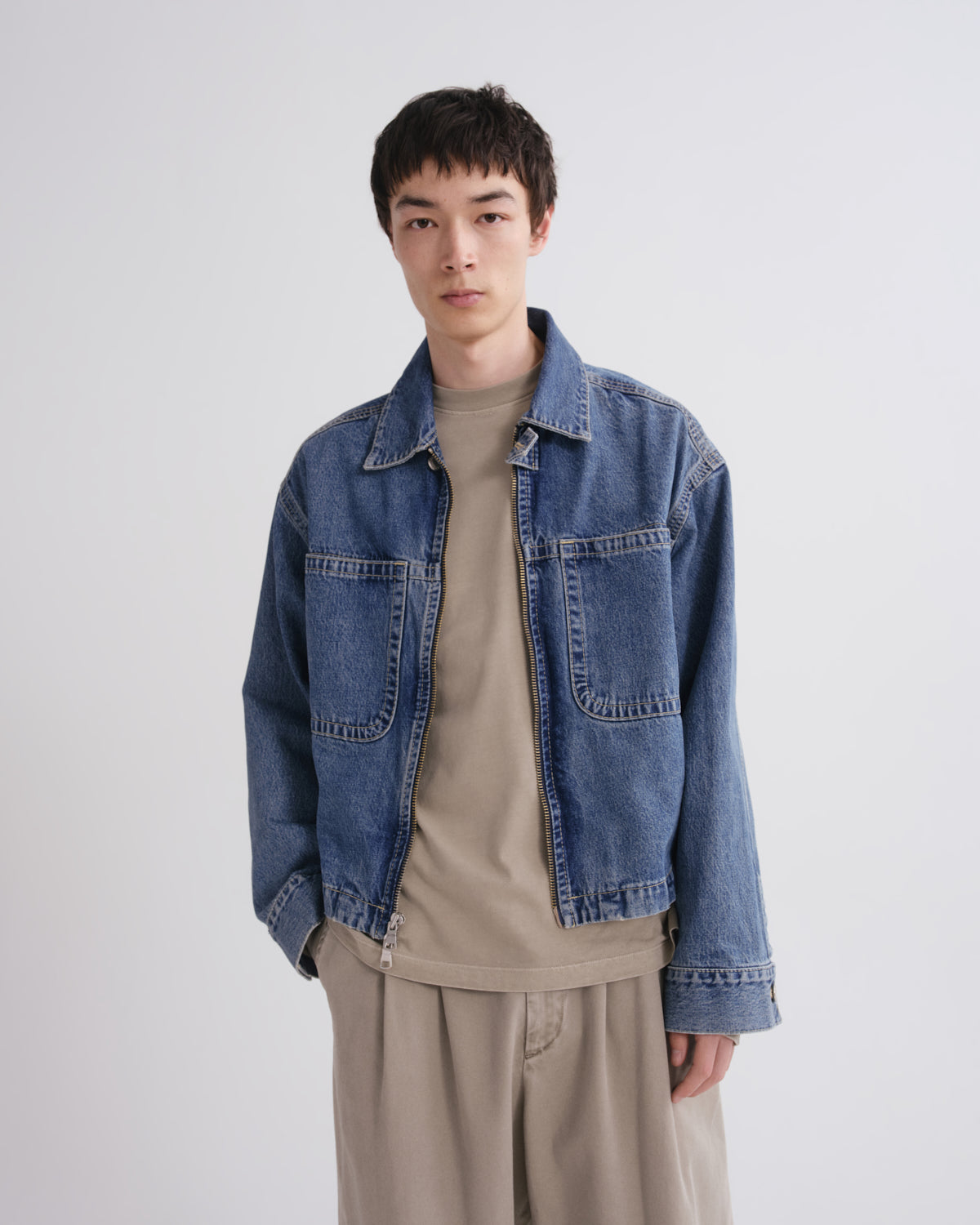 A model wearing a blue denim jacket over a beige t-shirt and beige trousers, standing against a light gray studio background.