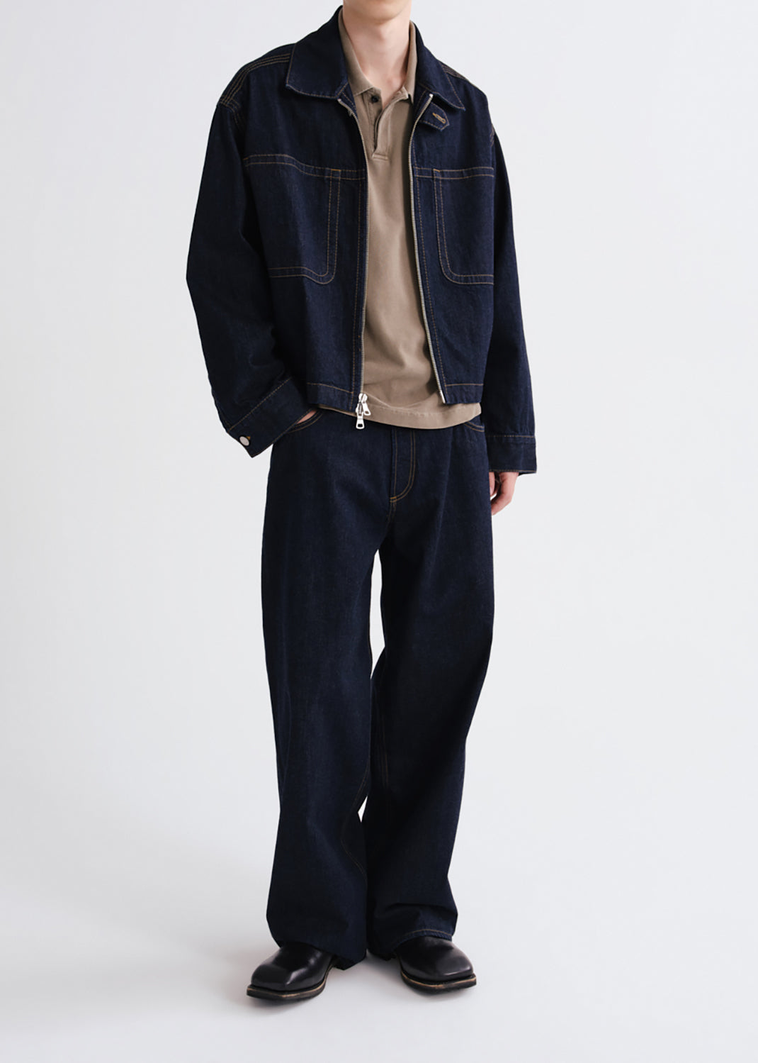 A model wearing a dark indigo denim jacket and matching wide-leg jeans, layered over a beige polo shirt and black leather shoes, photographed against a grey background.