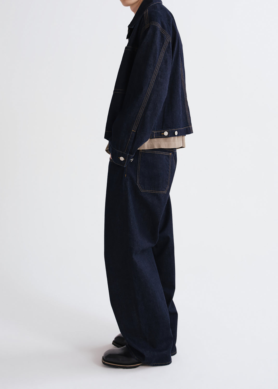A side profile view of a person wearing a dark navy blue denim jacket and matching wide-leg denim pants, styled with a beige shirt underneath and black shoes, against a grey background.