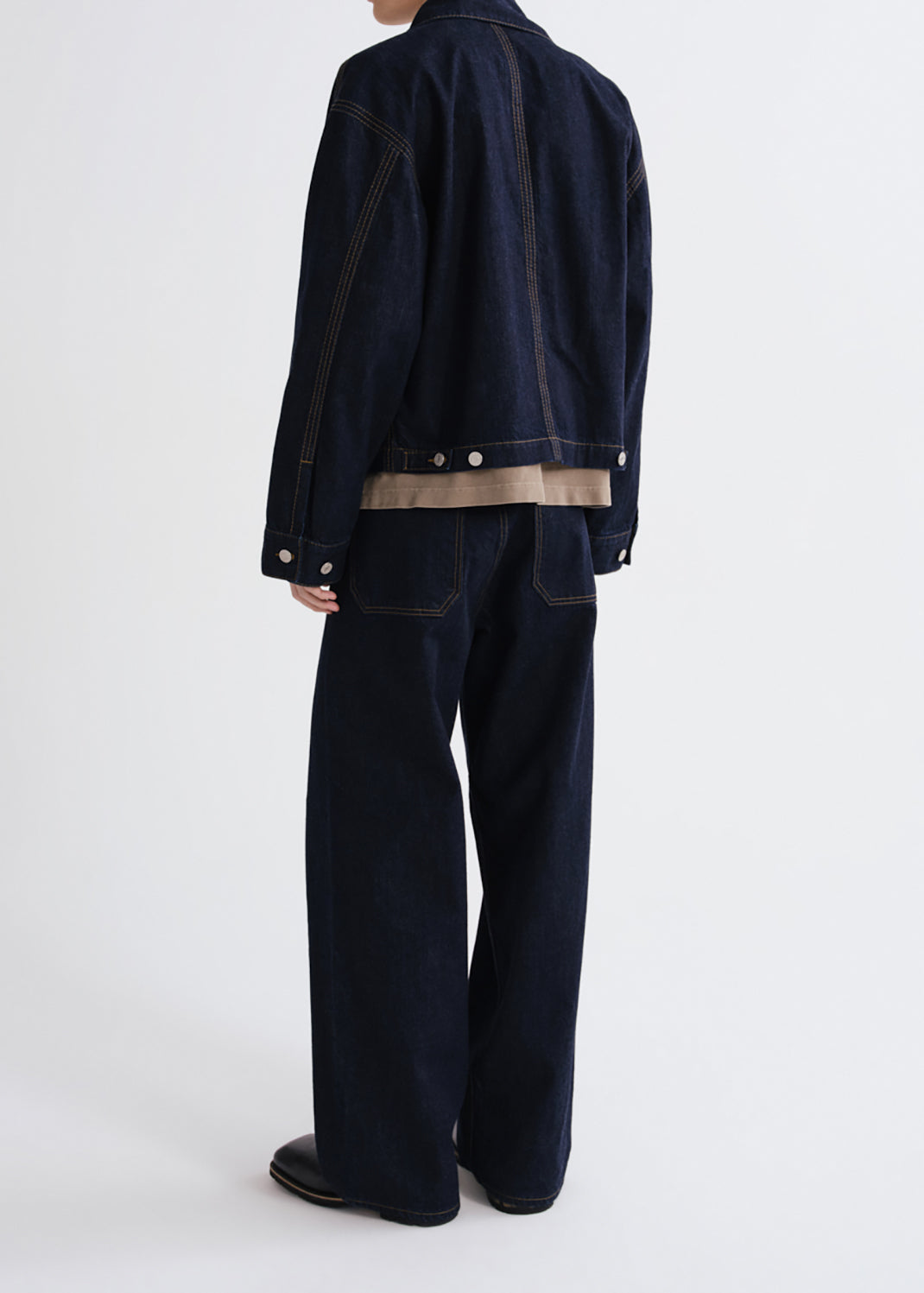 A model shown from the back wearing a dark indigo denim jacket and matching wide-leg jeans, featuring contrast tan stitching and a tan horizontal stripe detail near the hem.