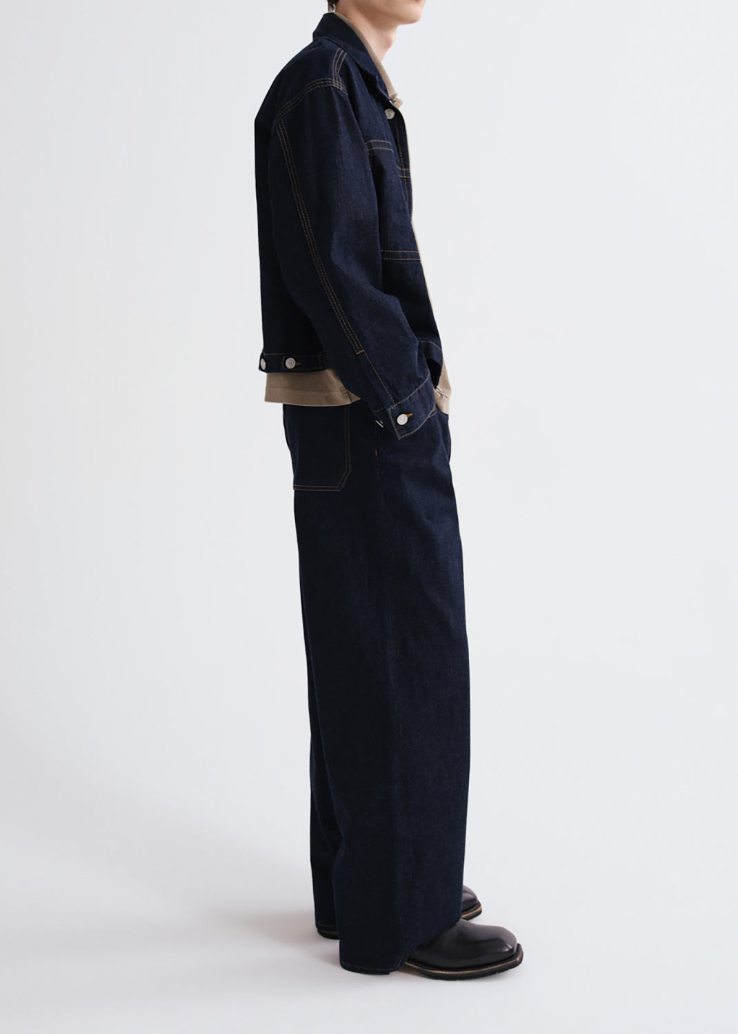 A model shown from the side wearing a dark navy blue denim jacket and matching wide-leg denim pants, styled with a beige shirt underneath and black leather shoes, against a grey background.