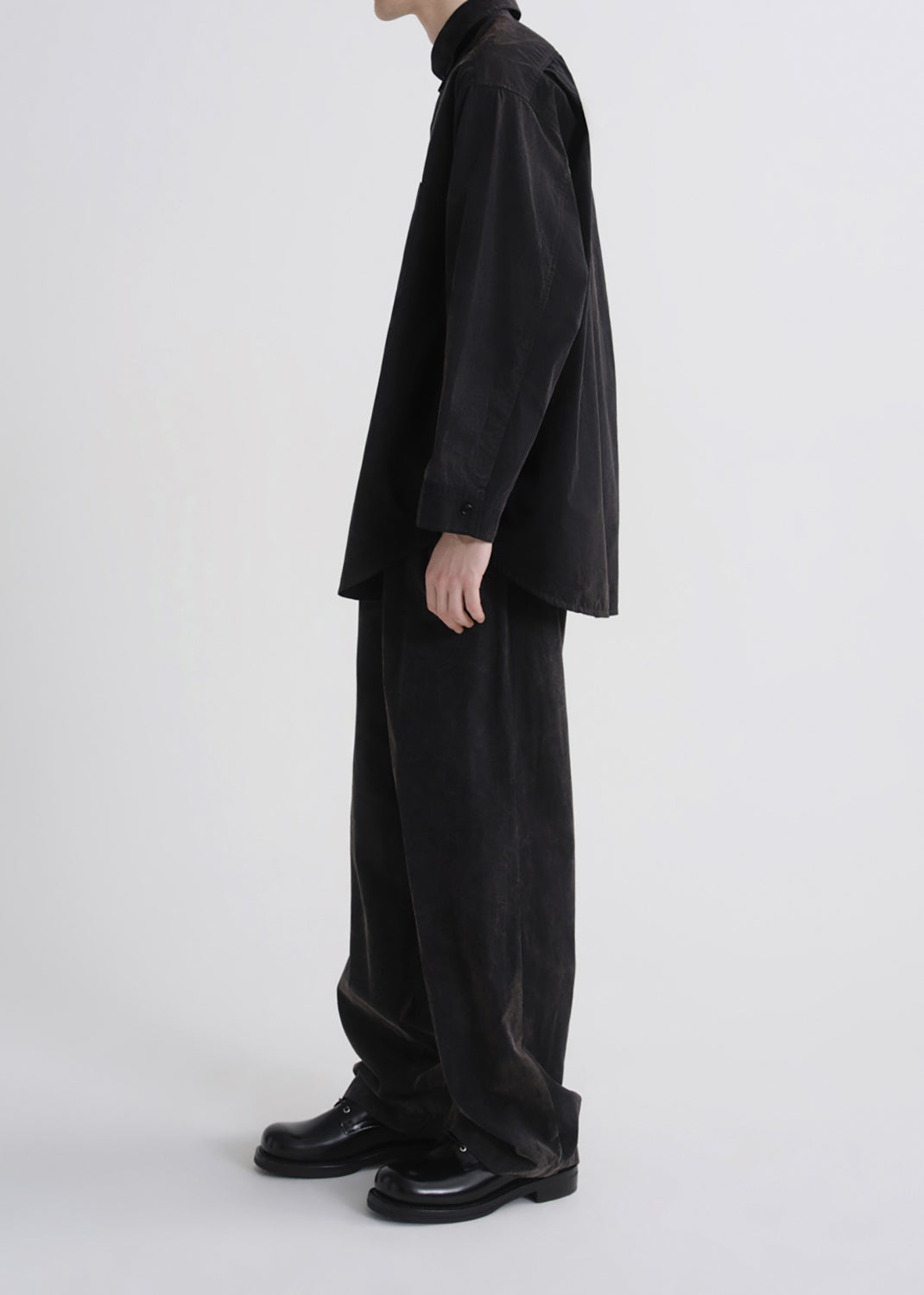 A model shown from the side wearing a black long-sleeve button-up shirt, black wide-leg corduroy trousers, and black leather shoes against a grey background.