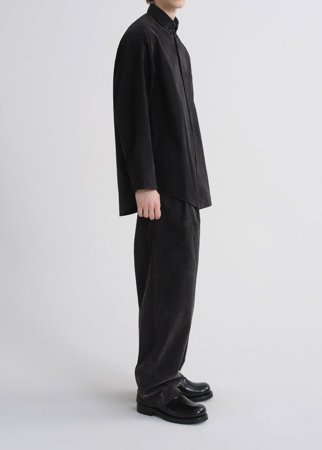 A model shown in profile wearing a black long-sleeve button-up shirt, black wide-leg trousers, and black leather loafers against a grey background.