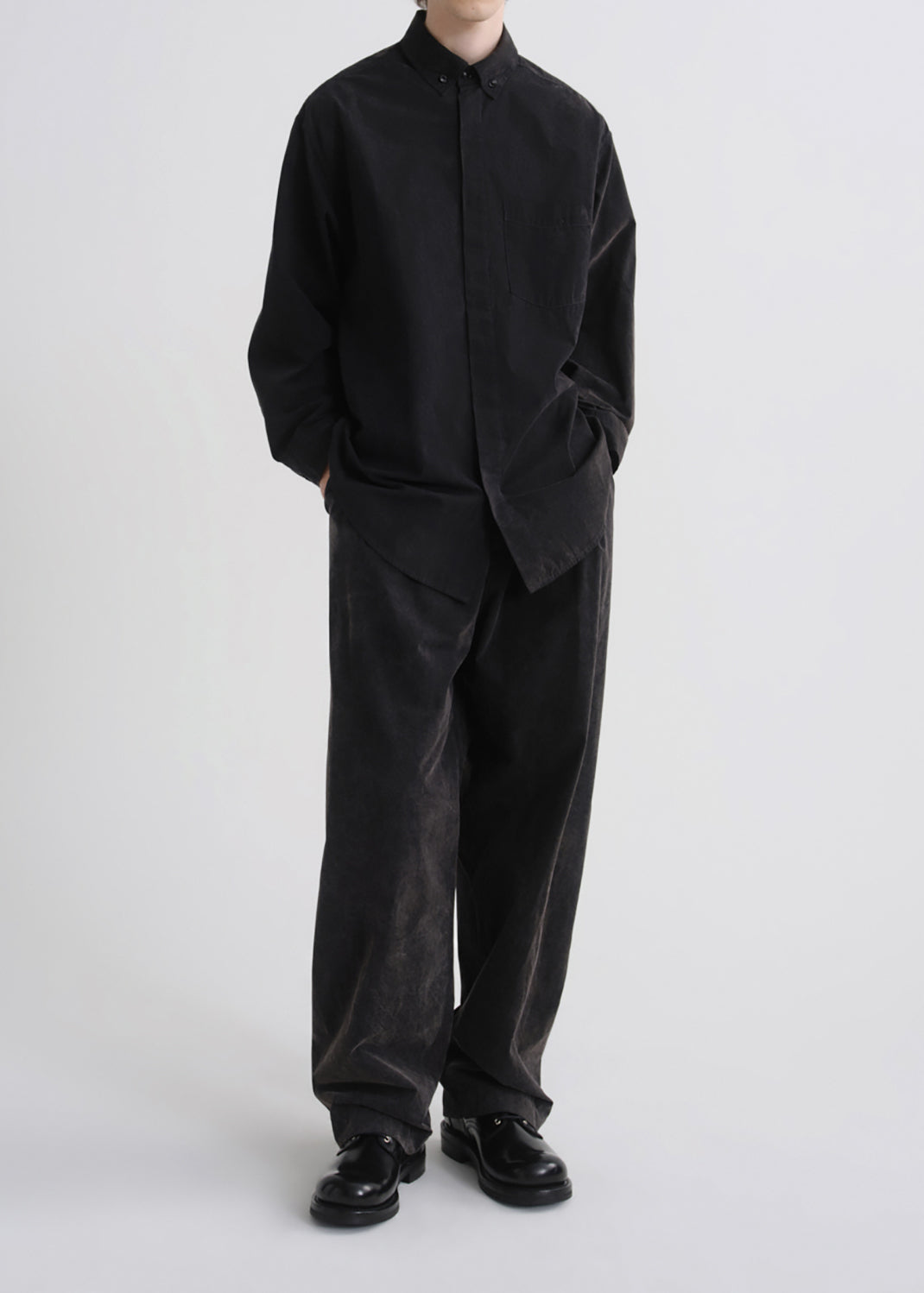 A model wearing a black long-sleeve button-up shirt and matching black corduroy trousers, paired with black leather shoes, against a grey background.