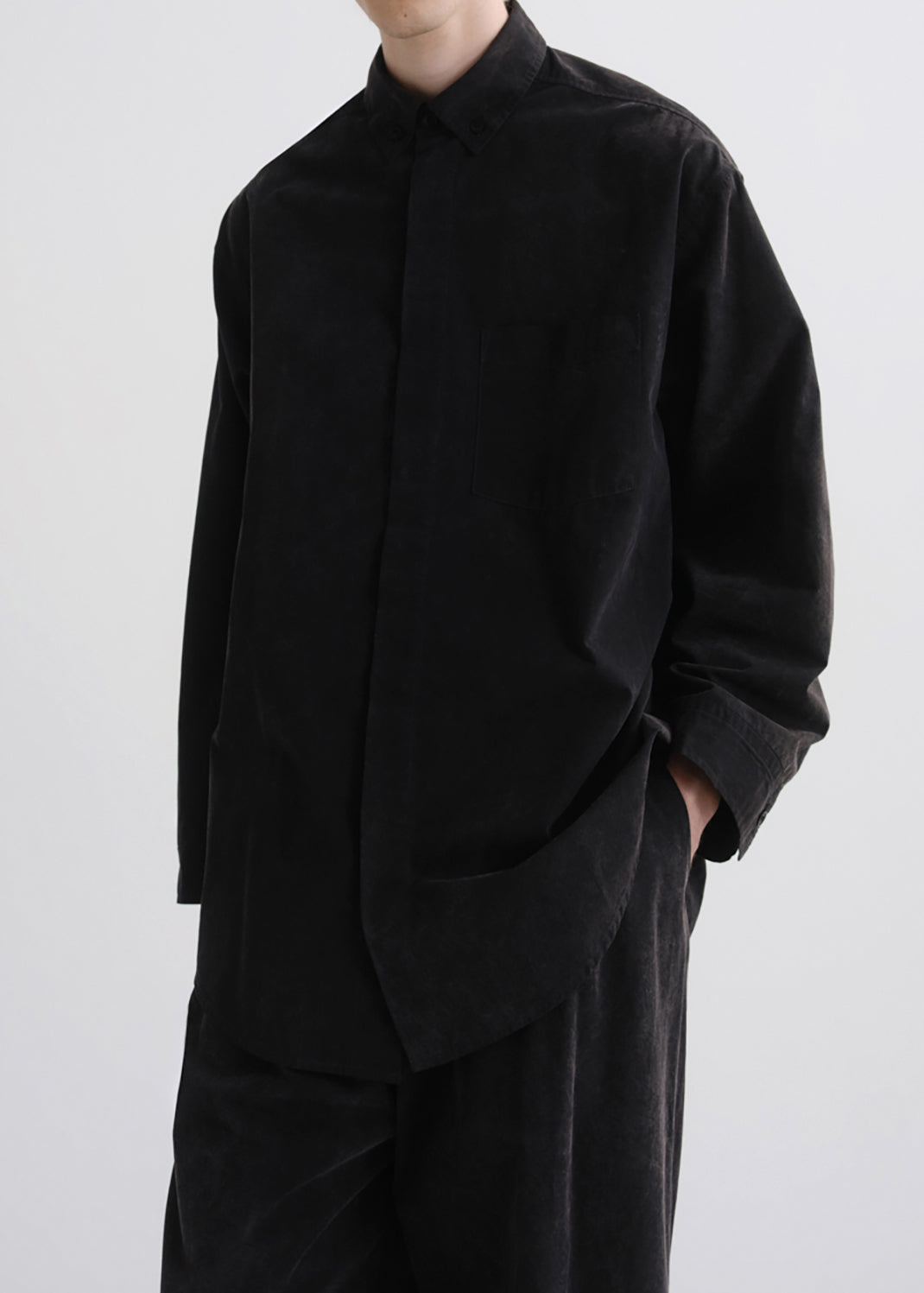 A person wearing an oversized black long-sleeve button-front shirt with a stand collar and matching black trousers, photographed against a grey background.