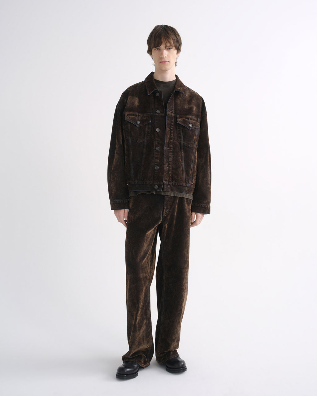 Person wearing a dark brown denim jacket with matching dark brown pants and black shoes on a grey background