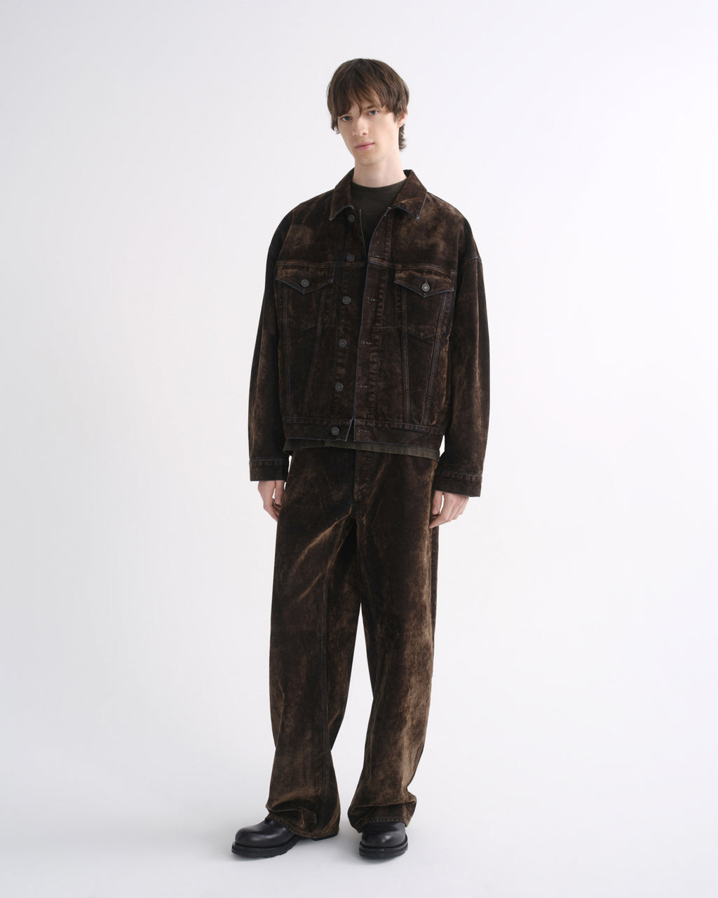 Person wearing a dark brown denim jacket with matching dark brown pants and black shoes on a grey background