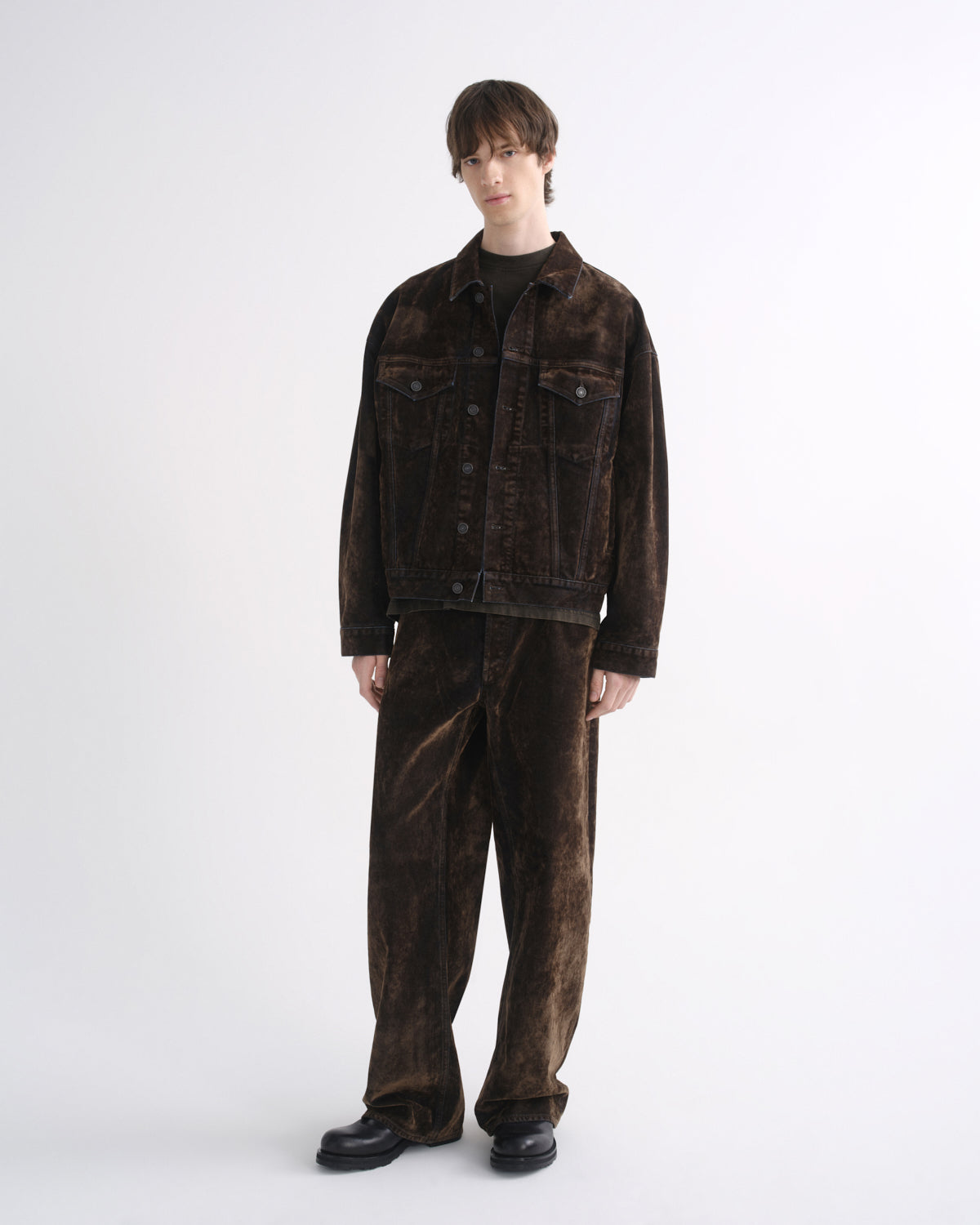 Person wearing a dark brown denim jacket with matching dark brown pants and black shoes on a grey background