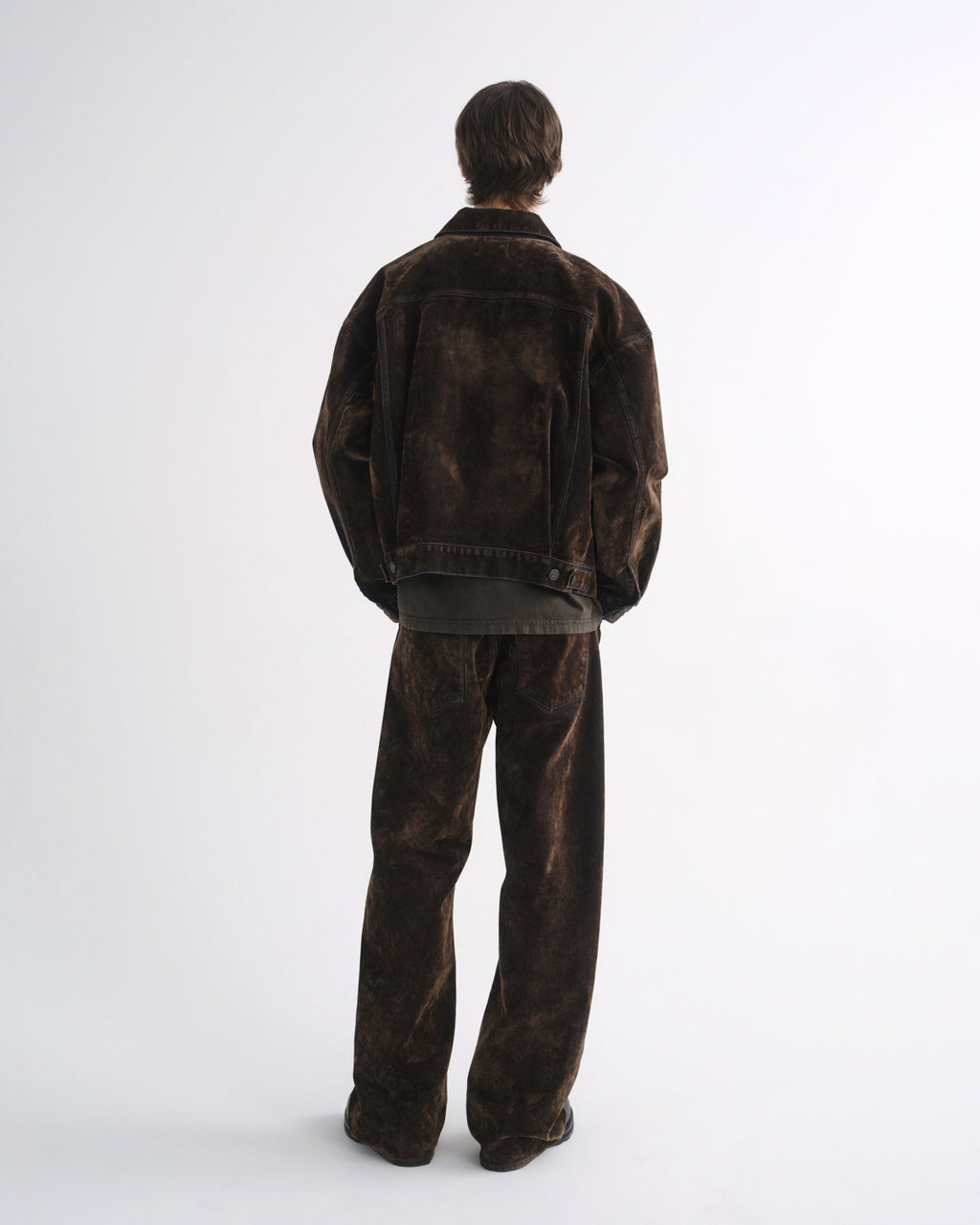 Person wearing a dark brown denim jacket with matching dark brown pants and black shoes, back view, on a grey background