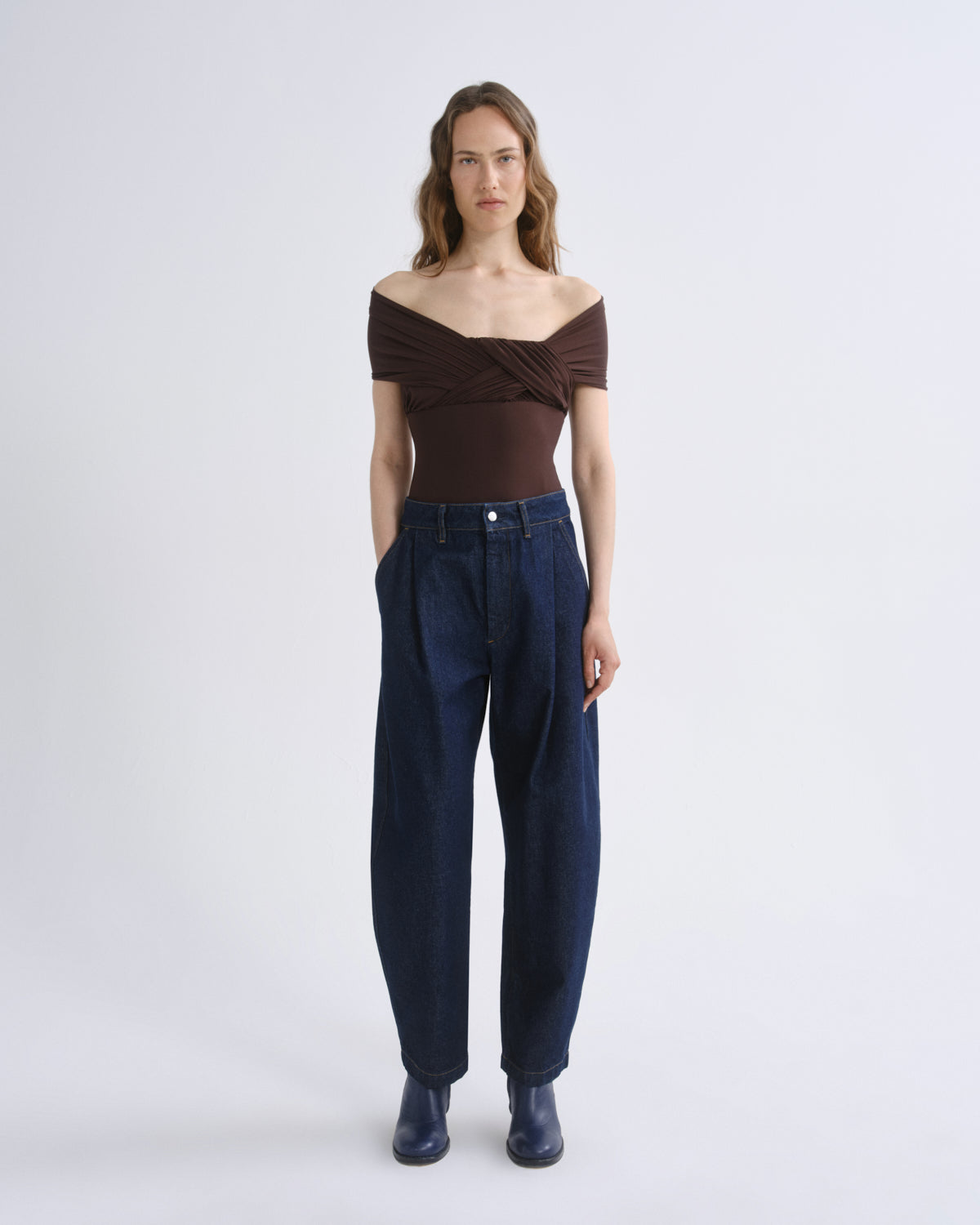 A model stands against a light gray studio background wearing a dark brown off-the-shoulder wrap top, dark blue wide-leg jeans, and dark blue boots.