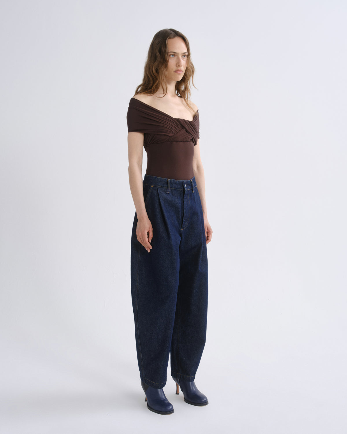 A model stands against a light gray studio background wearing a dark brown off-the-shoulder top, dark blue wide-leg jeans, and dark blue heeled boots.
