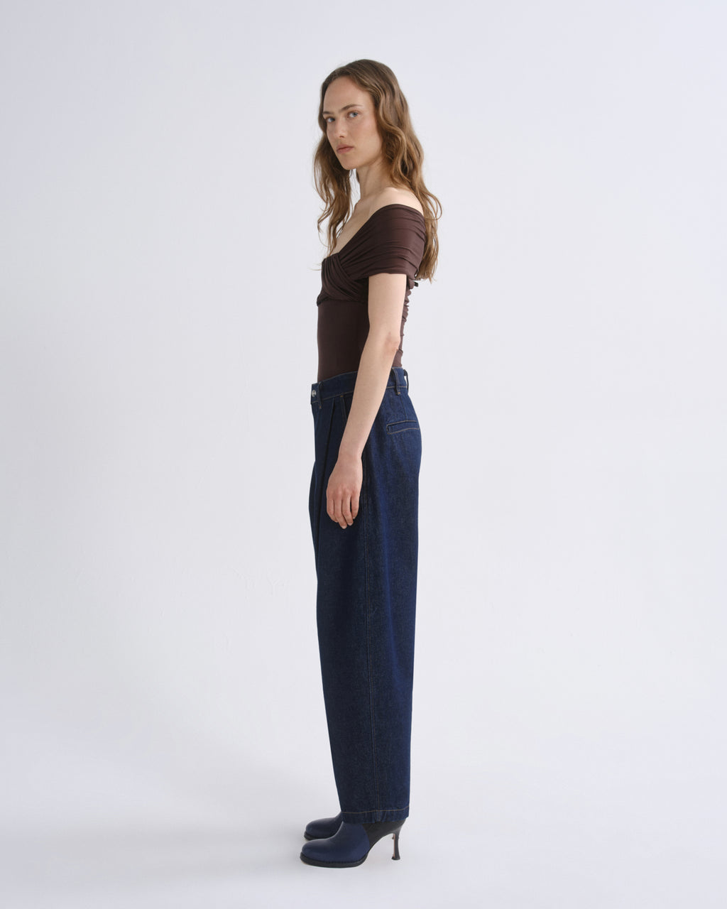 A model stands in profile wearing a dark brown off-the-shoulder short-sleeve top, dark blue wide-leg jeans, and dark blue heeled shoes against a plain light gray studio background.