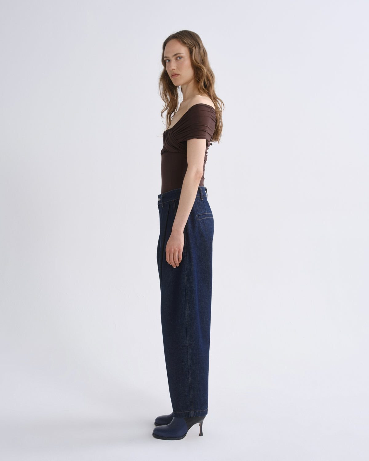A model stands in profile wearing a dark brown off-the-shoulder short-sleeve top, dark blue wide-leg jeans, and dark blue heeled shoes against a plain light gray studio background.