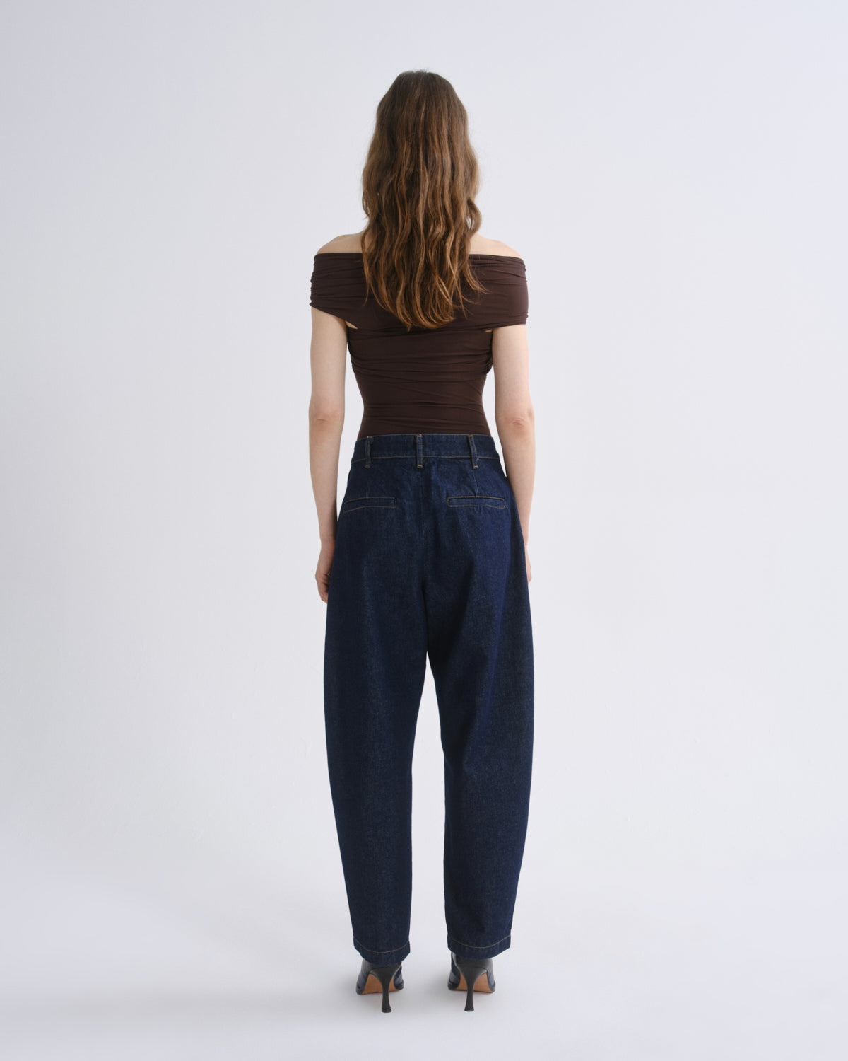 A woman viewed from the back wearing a dark brown off-the-shoulder top and dark blue wide-leg jeans, standing against a light gray studio background.