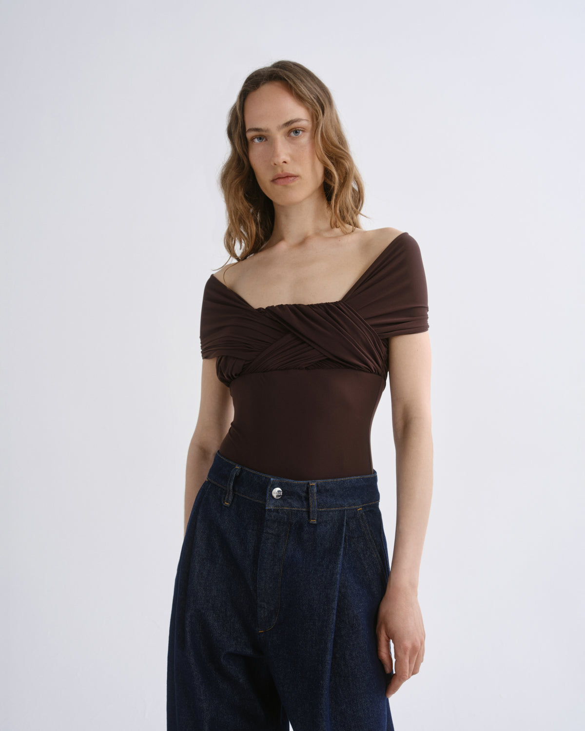 A woman modeling a dark brown off-the-shoulder top with a twisted front detail, paired with dark blue jeans, against a plain light gray studio background.