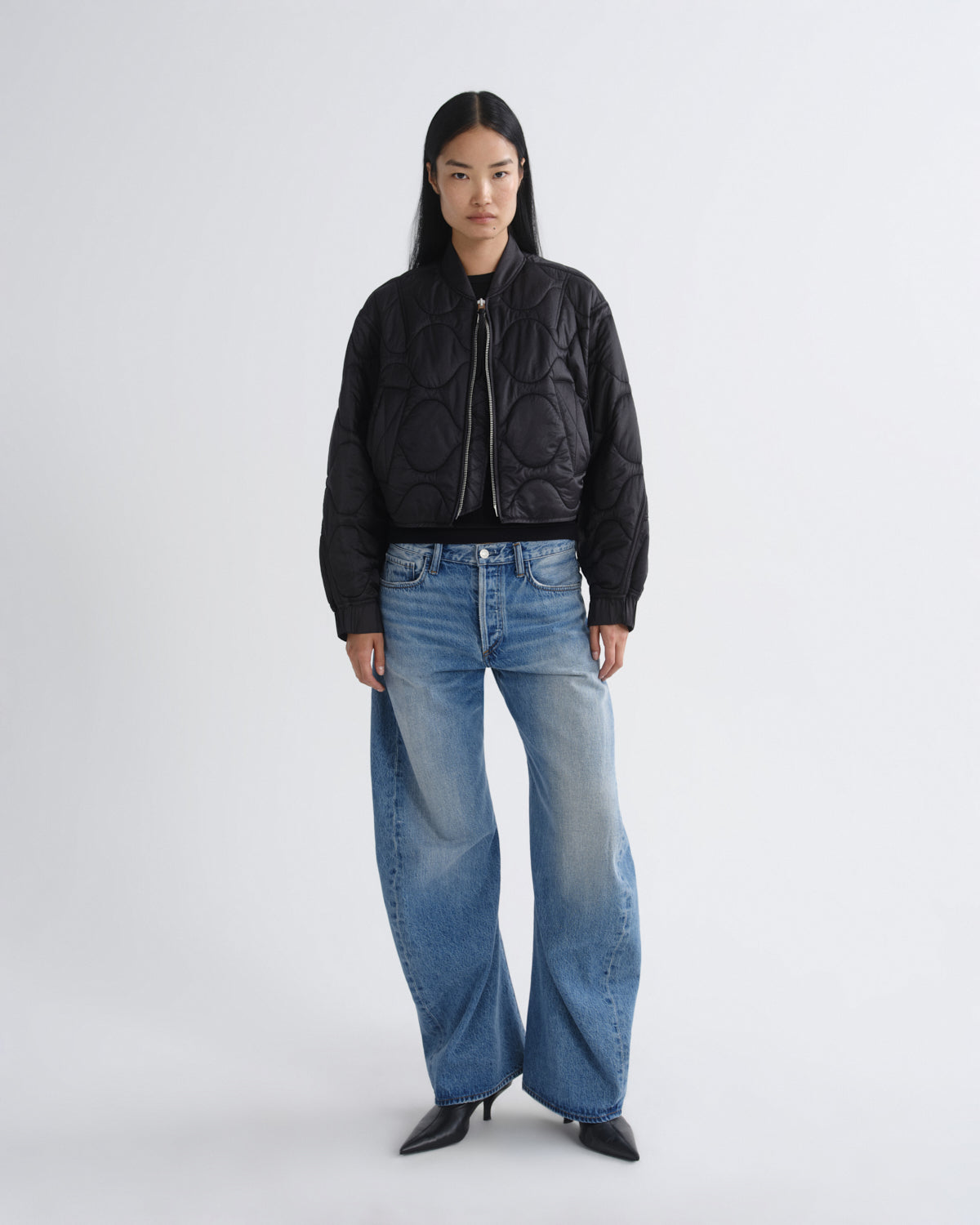 A model stands against a light gray studio background wearing a black quilted bomber jacket, wide-leg blue jeans, and black pointed-toe heels.