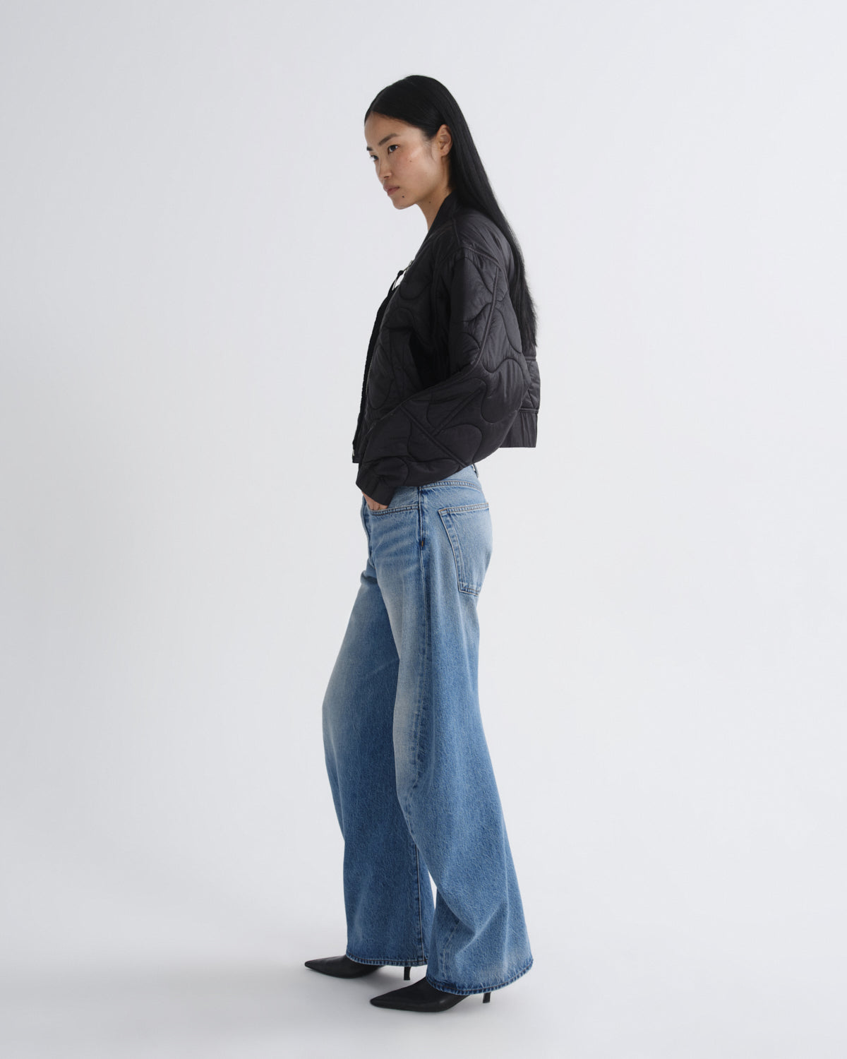 A woman stands in profile wearing a black quilted cropped jacket, wide-leg light blue jeans, and black pointed-toe heels against a plain light gray studio background.