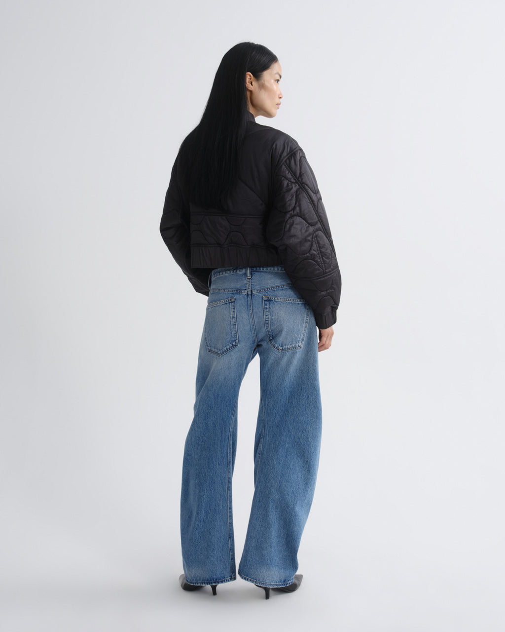 A model shown from the back wearing a black quilted cropped puffer jacket and wide-leg blue jeans, standing against a light gray studio background.