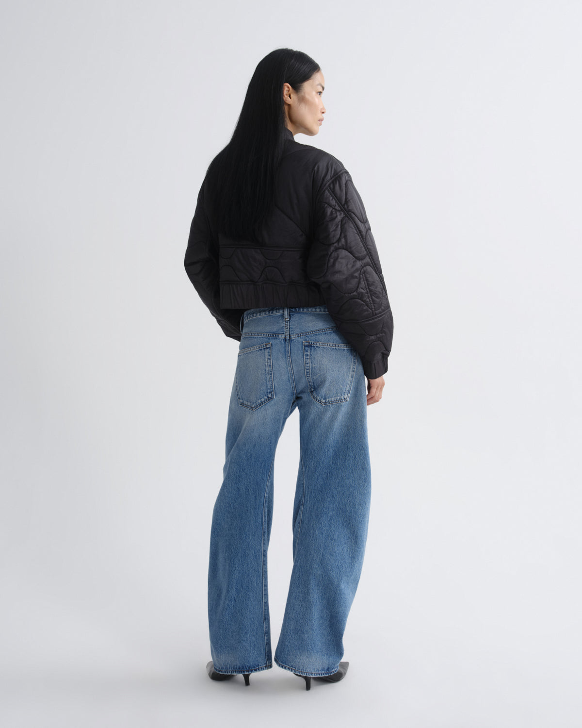 A model shown from the back wearing a black quilted cropped puffer jacket and wide-leg blue jeans, standing against a light gray studio background.