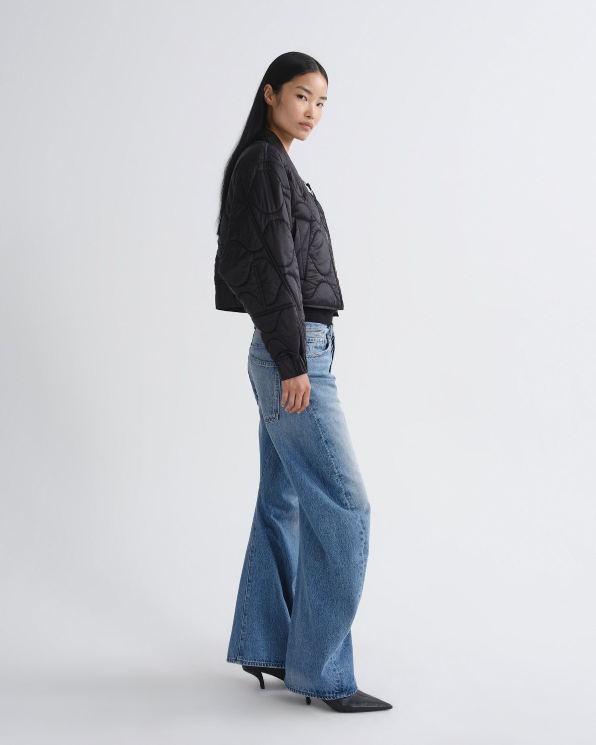 A woman modeling a black quilted puffer jacket paired with wide-leg blue jeans and black pointed-toe heels, standing in profile against a light gray studio background.