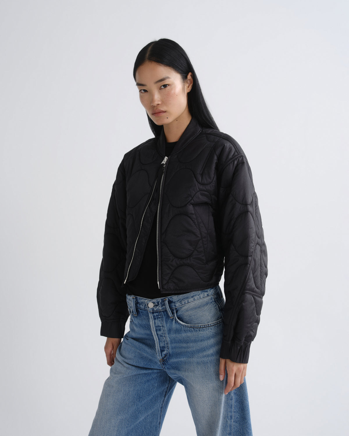 A model wearing a black quilted bomber jacket with silver zippers, paired with blue jeans, against a plain light gray studio background.